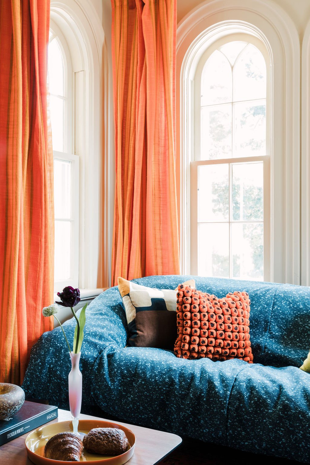 Sunlit living room with tall arched windows, orange curtains, a blue speckled sofa with patterned pillows, a small table with a vase and flowers, and a tray with baked goods.