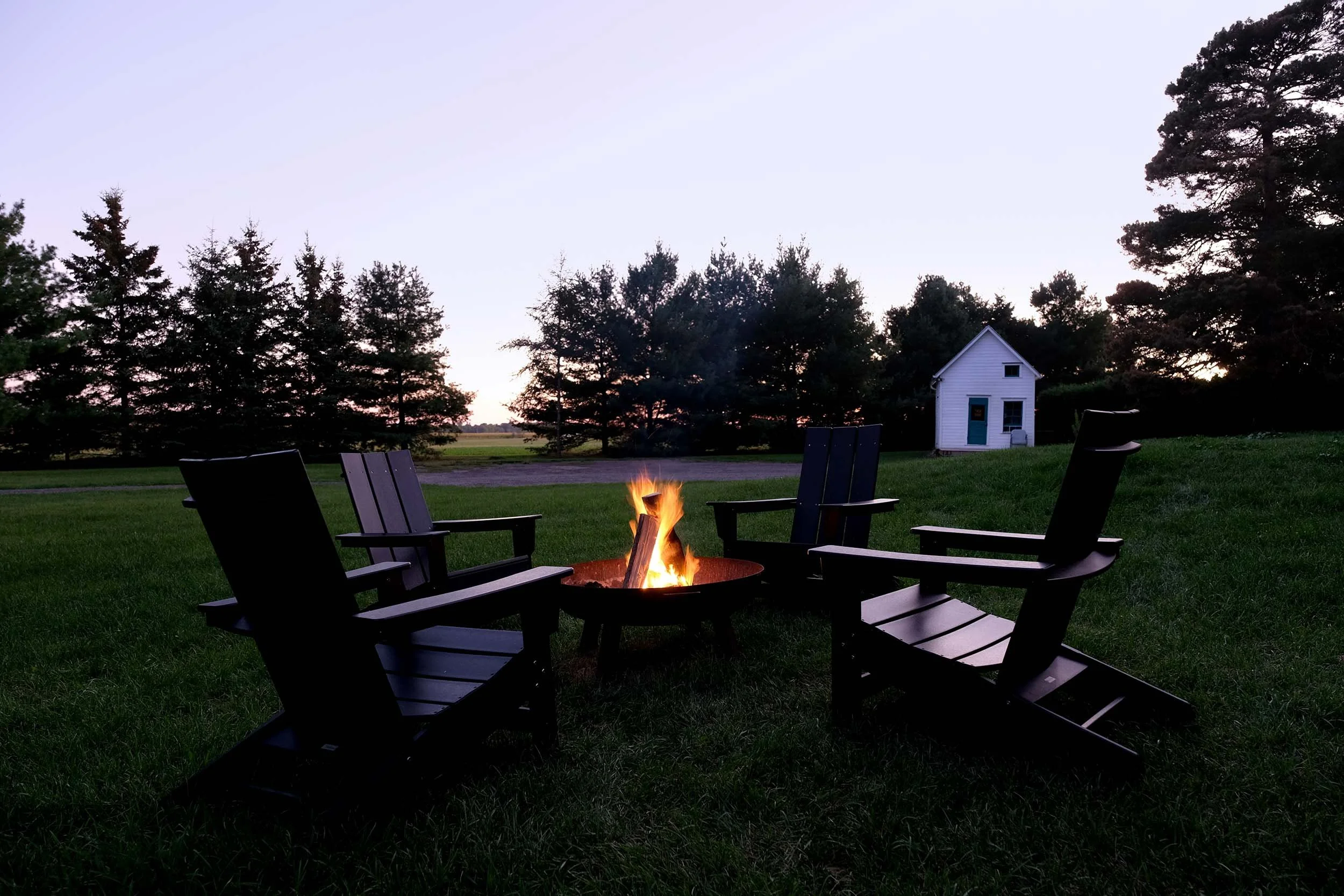 A backyard with a fire pit surrounded by chairs, a small white house, and tall trees in the background during dusk or early evening.
