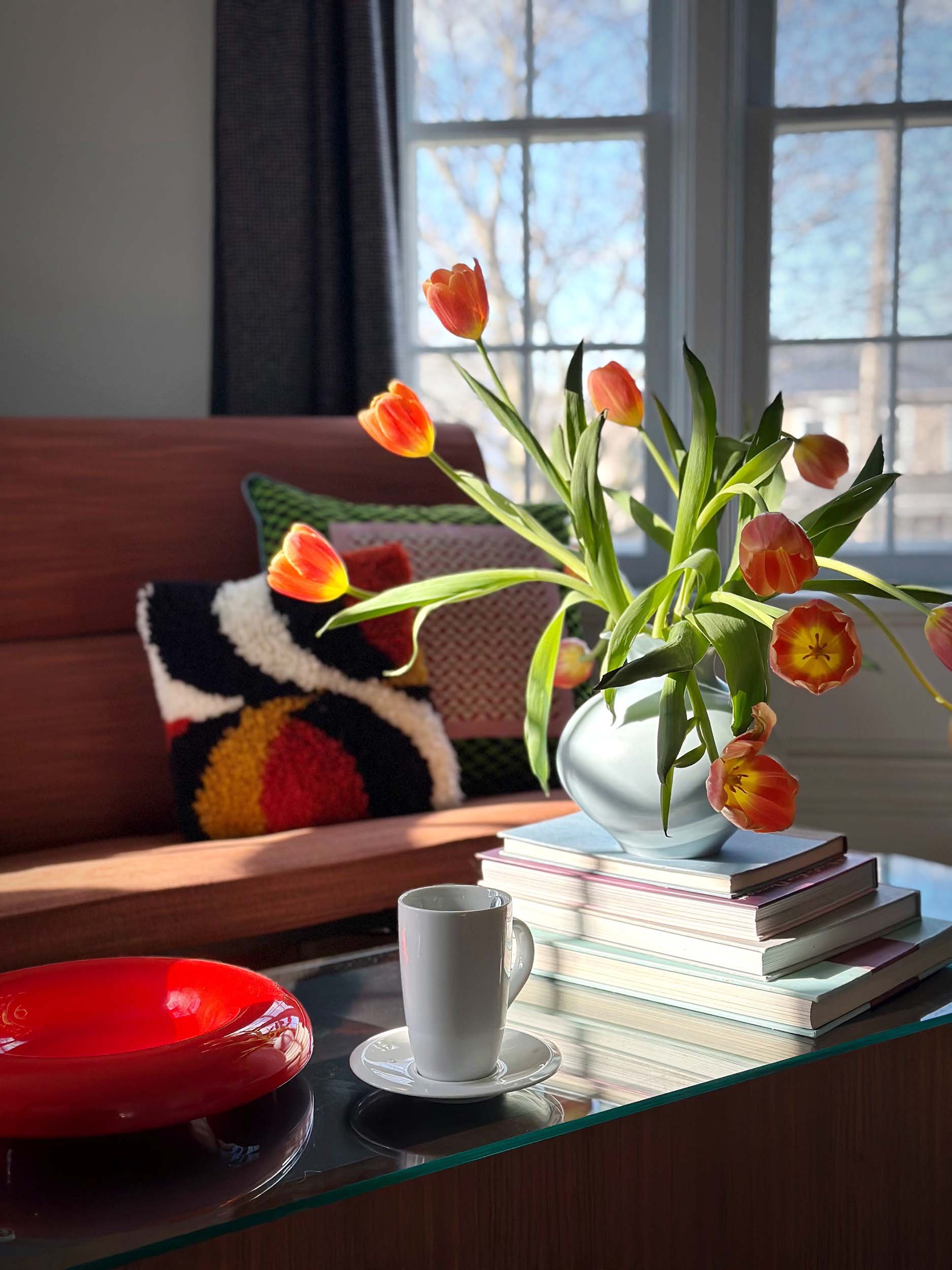 A vase of orange tulips on top of a stack of books on a glass table, with sunlight streaming through a window in the background, a ceramic cup and a red plate nearby, and a colorful knitted pillow on a wooden couch.
