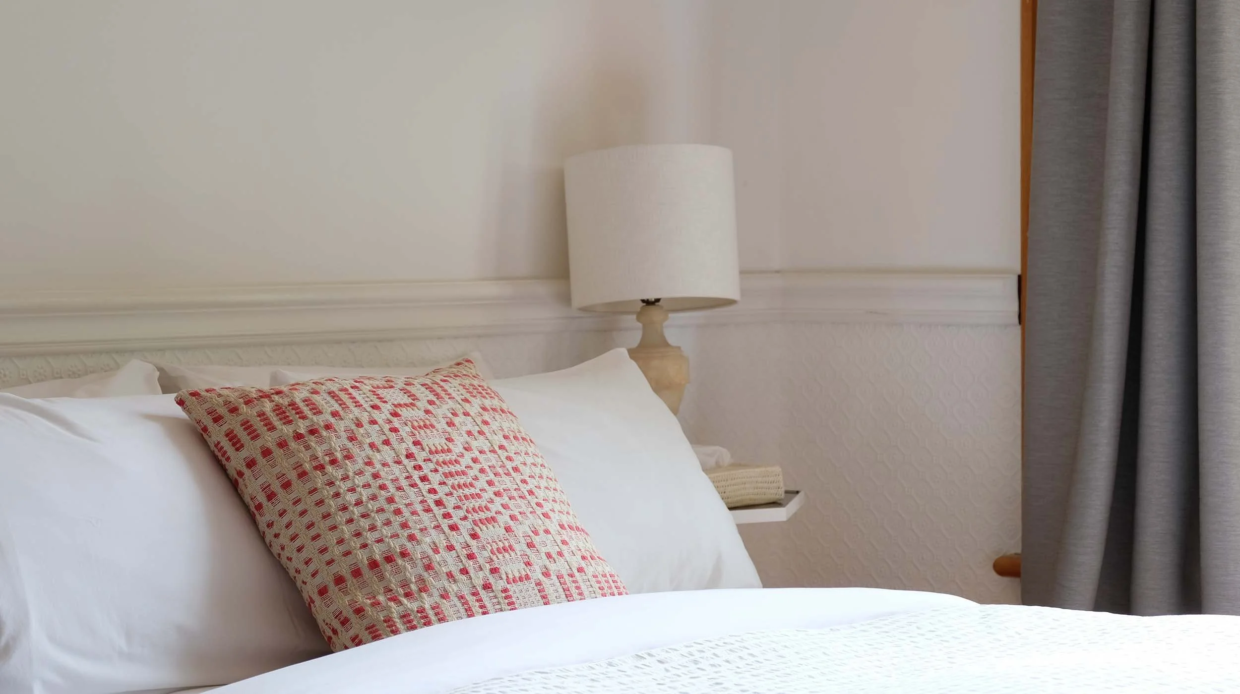 A neatly made bed with white sheets, a decorative red and beige patterned pillow, a cream-colored lamp on a nightstand, and gray curtains on the side of the bed.