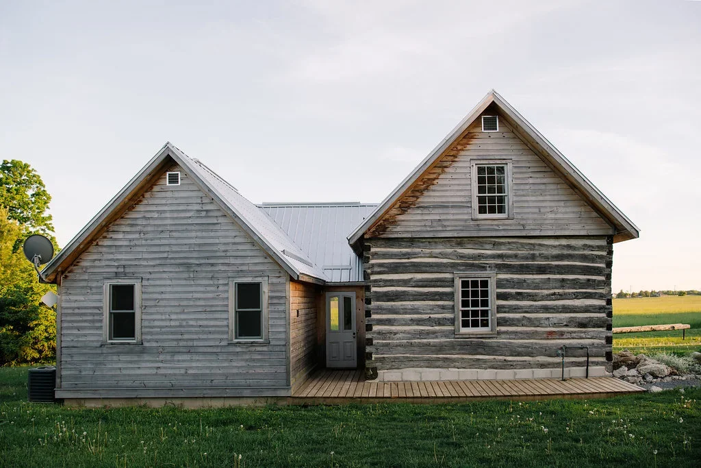 A rustic gray wooden house with two gable roofs, three visible windows, a small porch, and a grassy yard, set in a rural landscape.