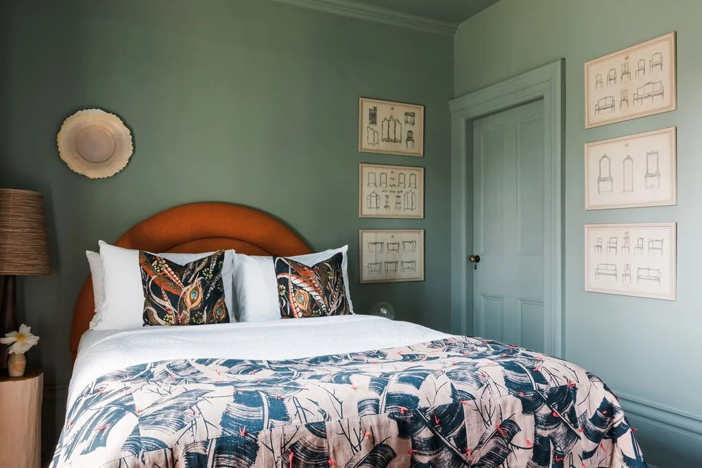 A bedroom with a neatly made bed featuring leaves and bird print blanket and decorative pillows, a nightstand with a flower in a pot, a wall-mounted hat, and framed sketches of chairs and furniture on the walls.