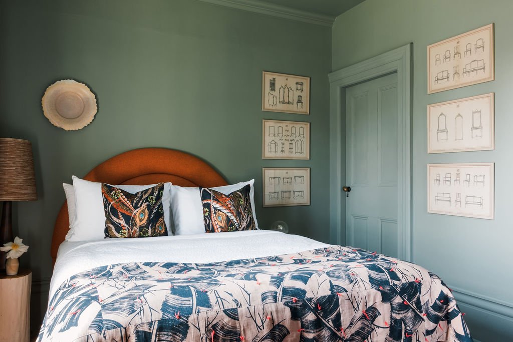 A neatly made bed in a bedroom with light green walls, with a brown curved headboard, two decorative pillows with peacock feather patterns, and a white bedspread with a tropical leaf design. To the left, there is a wooden nightstand with a lamp and a flower in a small vase. On the walls, there are framed sketches of various chairs and furniture.