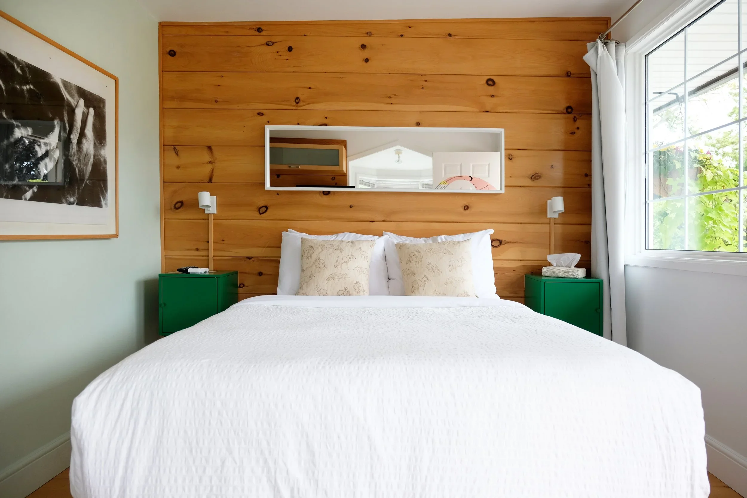 A cozy bedroom with a wooden accent wall, a large bed with white bedding, green nightstands, a framed black-and-white art piece, and a large window with white curtains showing greenery outside.
