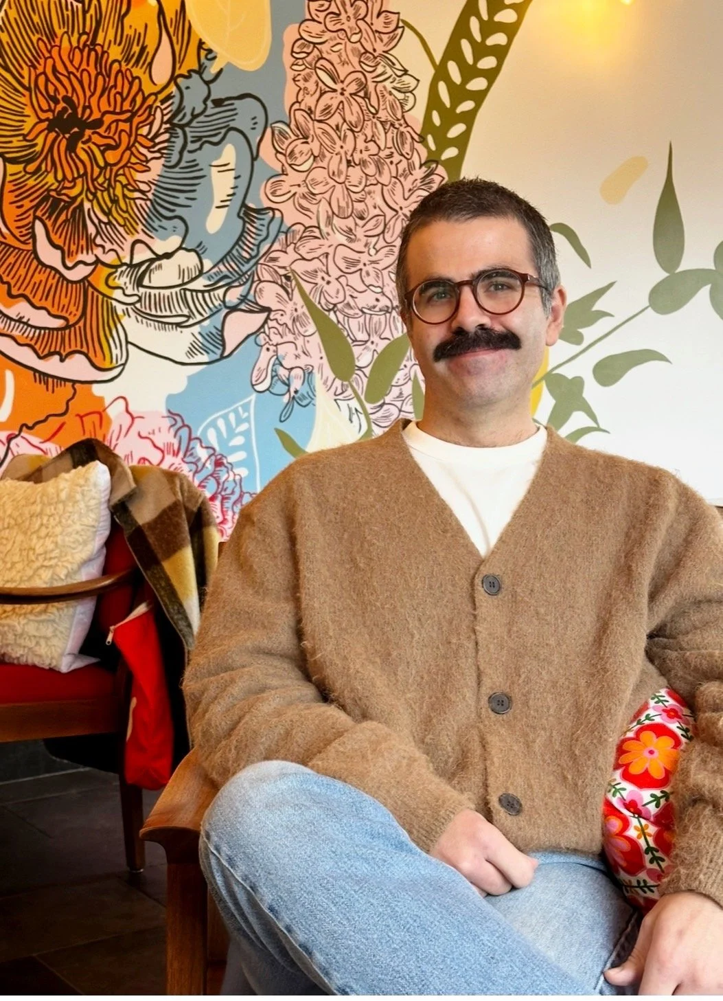 A man with glasses and a mustache wearing a brown cardigan and white t-shirt, sitting in a colorful floral mural background, with a bench and pillows to his left.