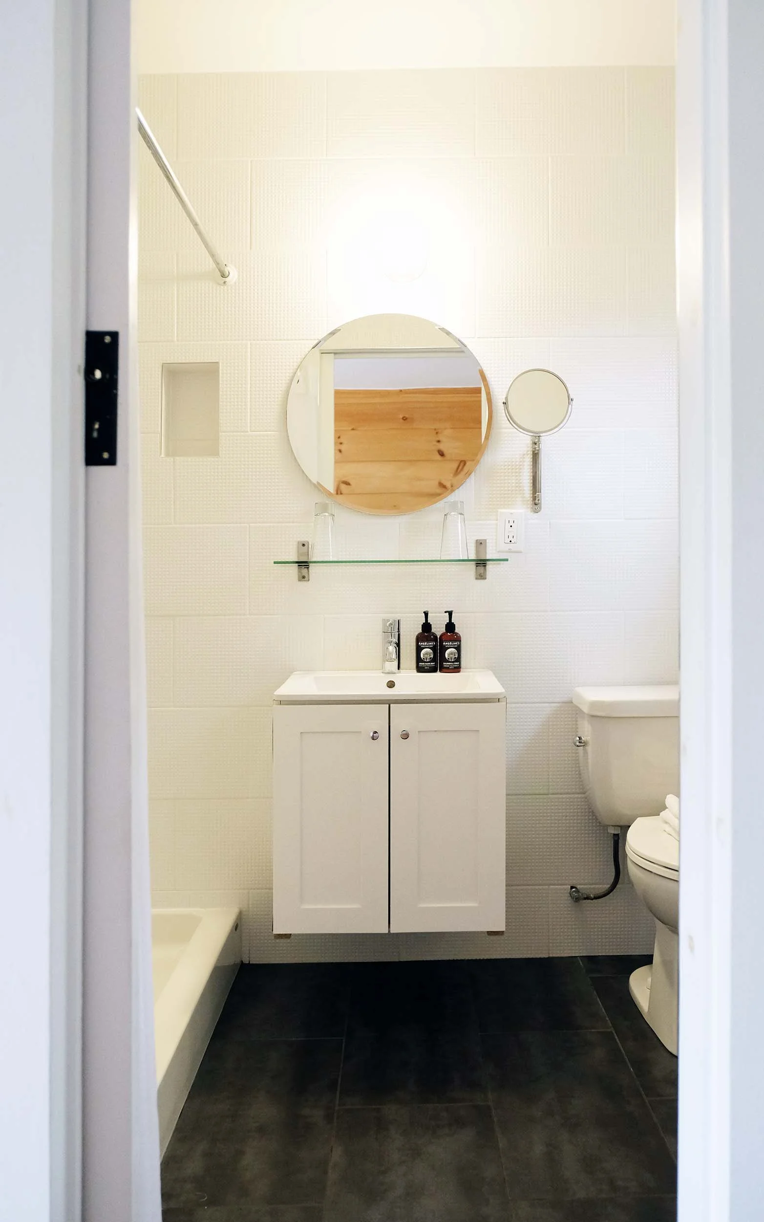 Small bathroom viewed from doorway, featuring a white vanity with a single sink, mirror, and two black soap dispensers. To the right, there is a toilet with a round seat and toilet paper. The walls are tiled off-white, and the floor is dark tile. A small built-in wall niche is on the left wall.