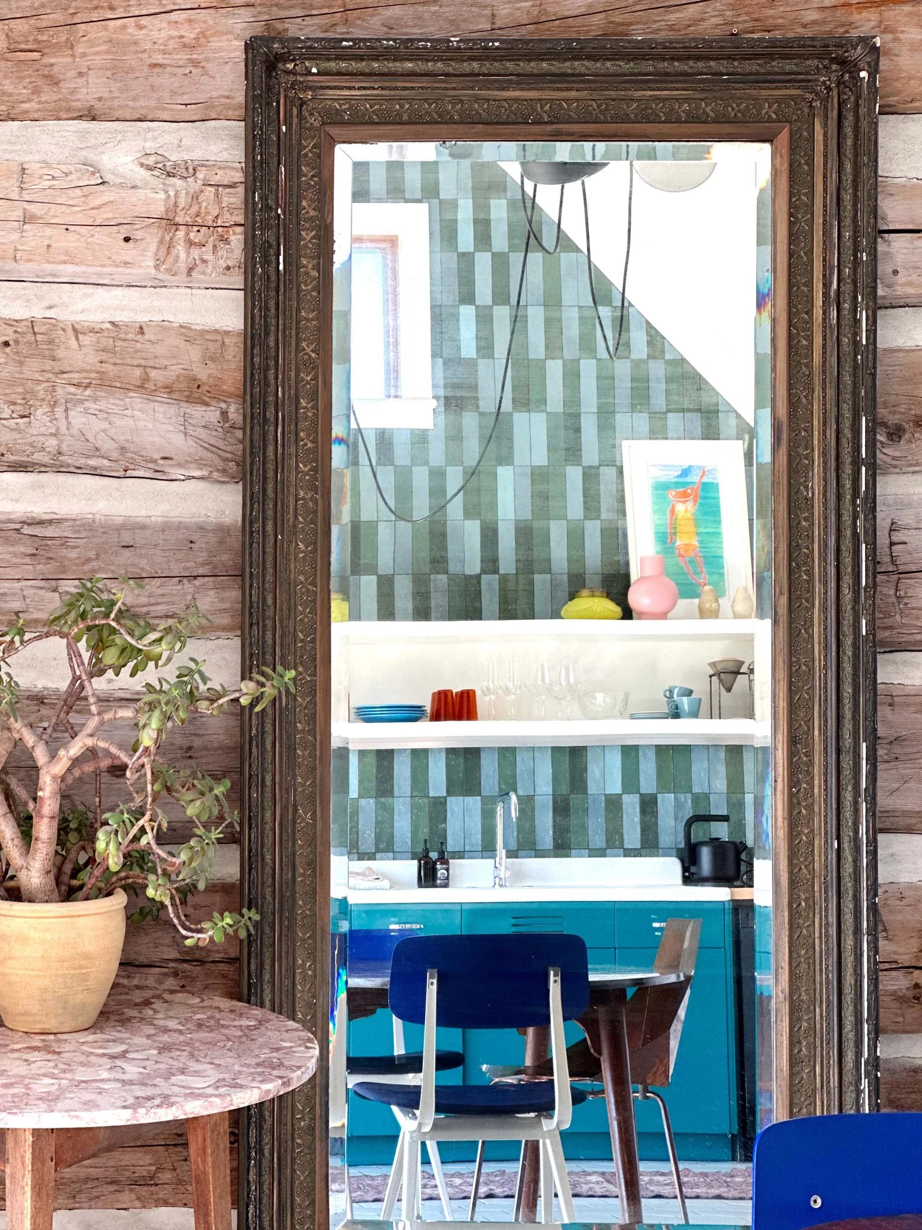 A reflection in a mirror shows a kitchen with green checkered tiles, a white shelf holding colorful vases and artwork, a blue cabinet, and a dining area with chairs. The mirror is mounted on a wooden wall, and a potted plant is seen on a table in the foreground.