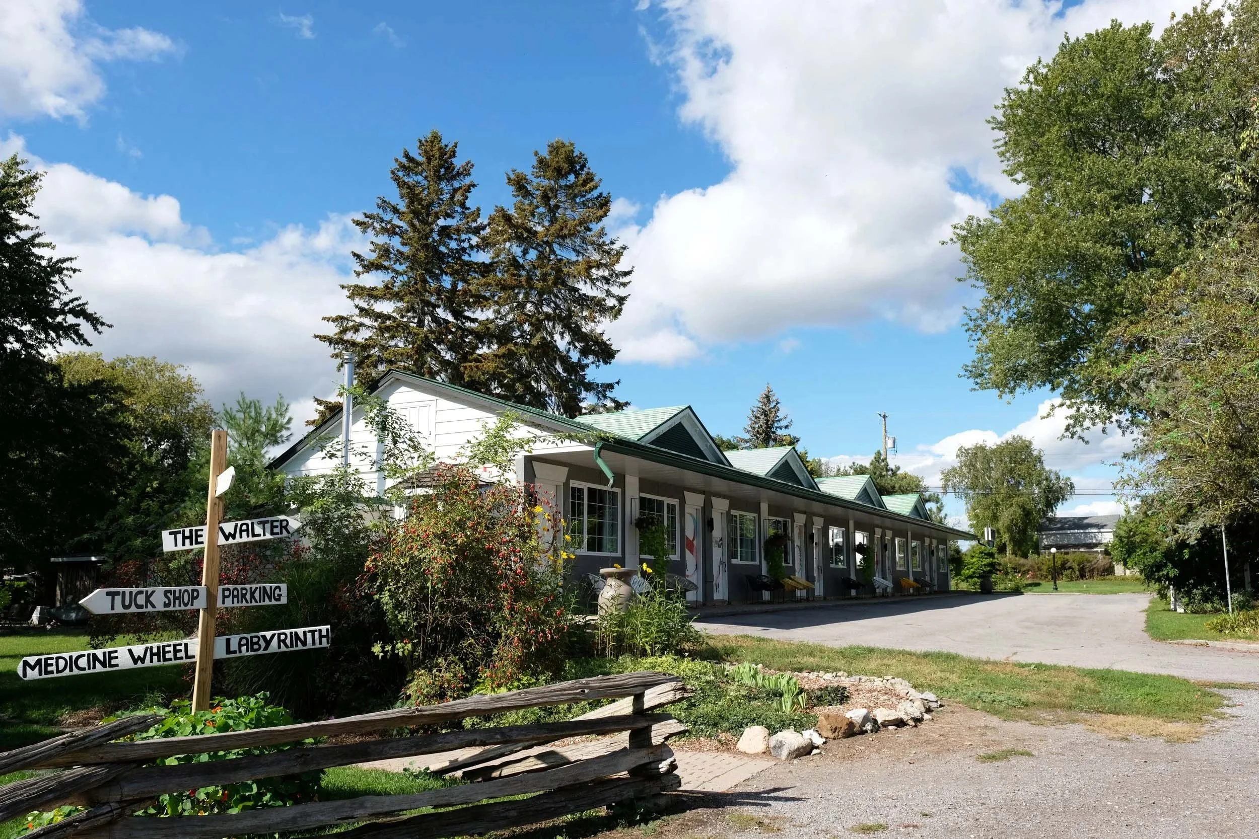 A white building with a green roof and a porch, surrounded by trees and shrubs. A signpost with multiple signs points to various locations. The sky is blue with some clouds.