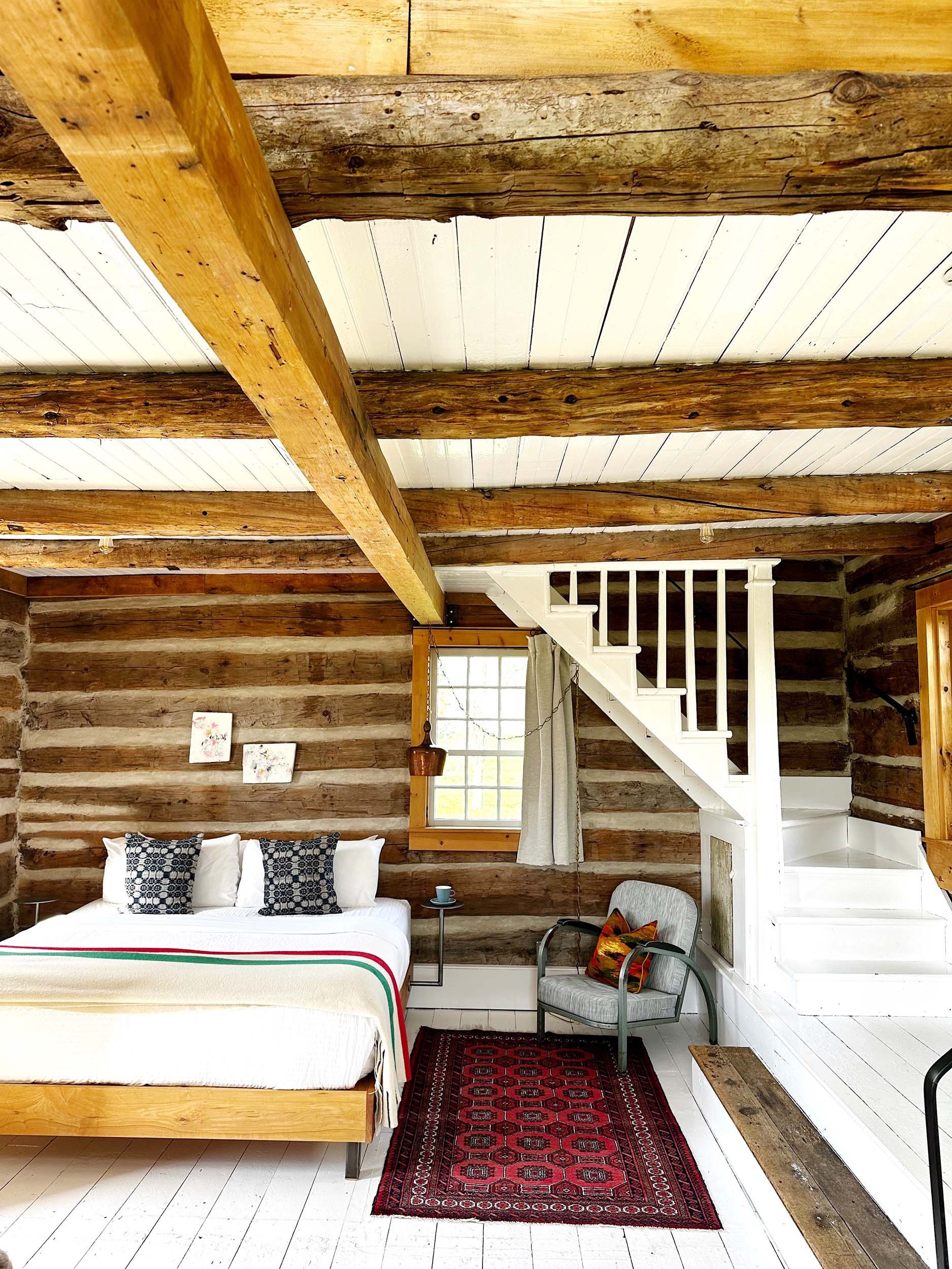 Cozy rustic bedroom with wooden log wall, white ceiling, bed with white bedding and black patterned pillows, red patterned rug, small side table with a cup, vintage armchair with colorful pillow, window with light curtains, staircase with white railing.