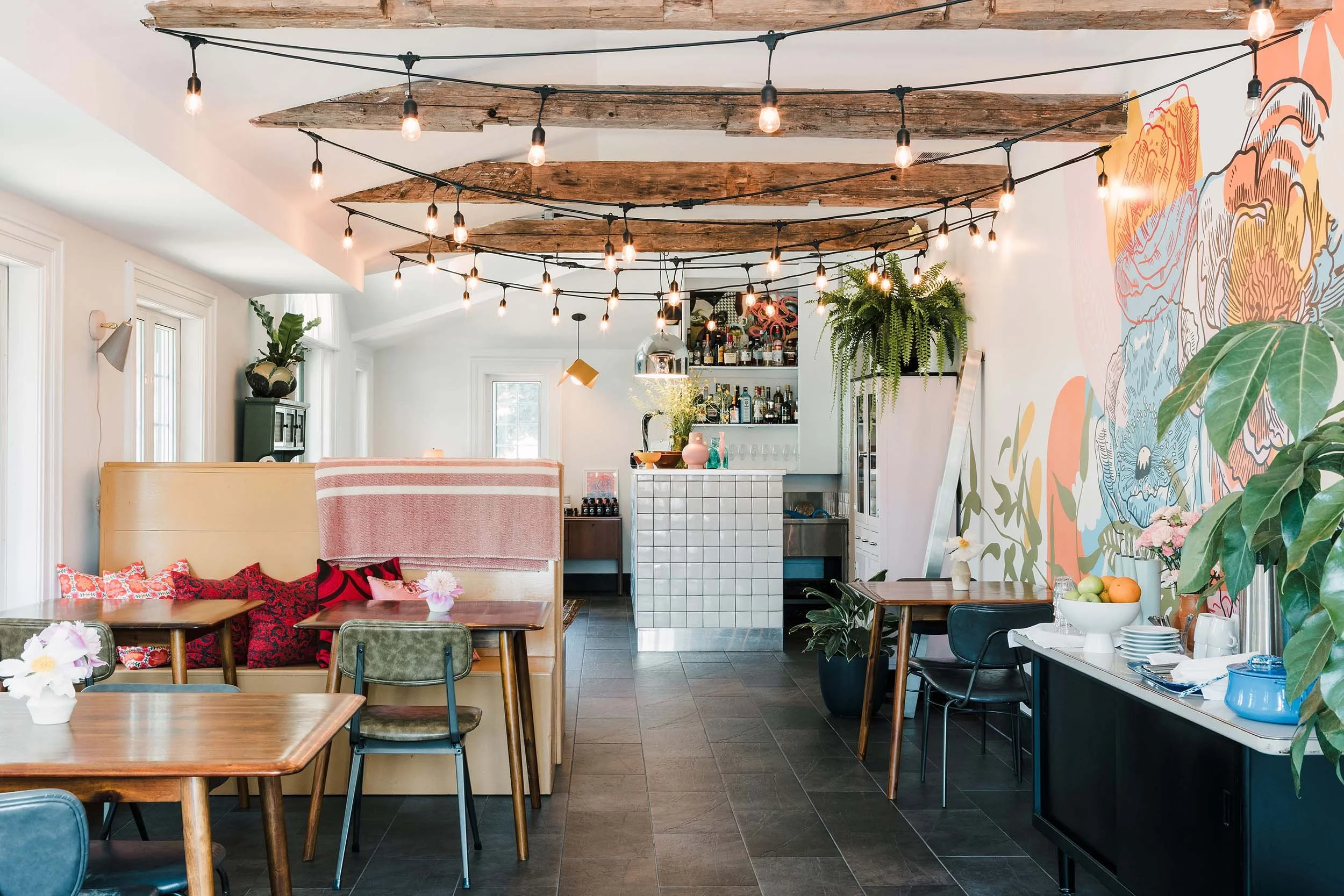 Interior view of a cozy cafe or restaurant with hanging string lights, wooden ceiling beams, eclectic furniture, a mural on the wall, a bar area with bottles, and decorative plants.
