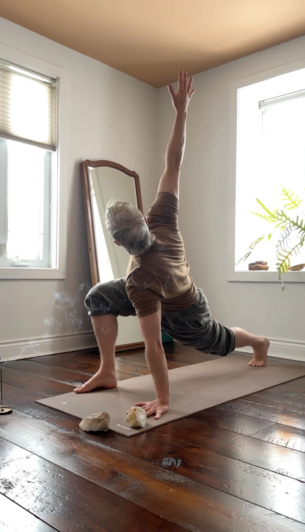 A person practicing yoga indoors on a mat, performing a one-arm downward dog pose with one arm reaching towards the ceiling and head tilted to the side, with windows and a mirror in the background.