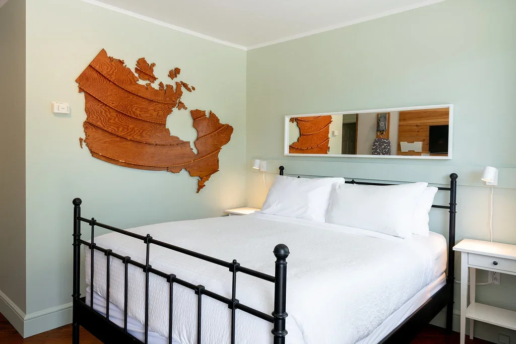 A bedroom with a black metal bed frame, white bedding, and two white nightstands with small lamps. A wooden map of North America decorates the pale green wall above the bed, and a rectangular mirror reflects part of the room.