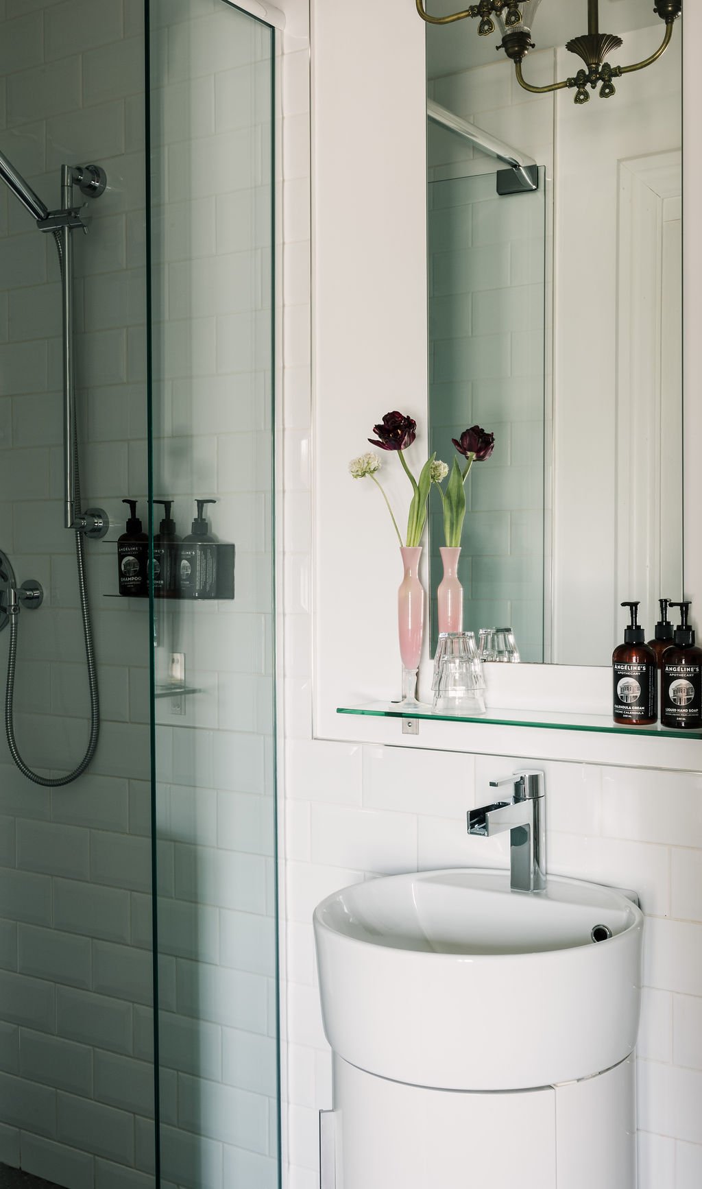 Bathroom with a glass shower, white tiled walls, a modern sink, a mirror, a glass shelf with pink vases and flowers, glasses, and bottles of hand soap.