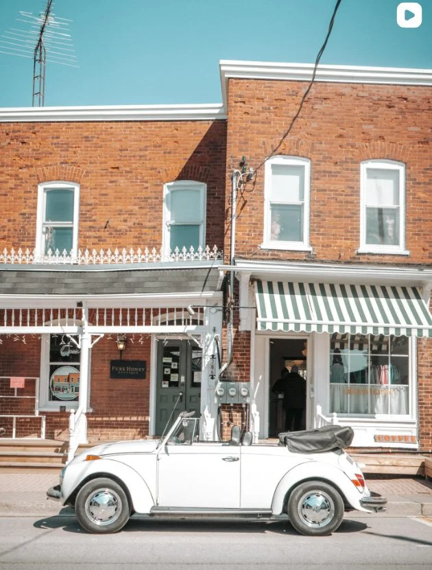 A white vintage convertible car parked on the street in front of a brick building with a striped awning and a storefront.