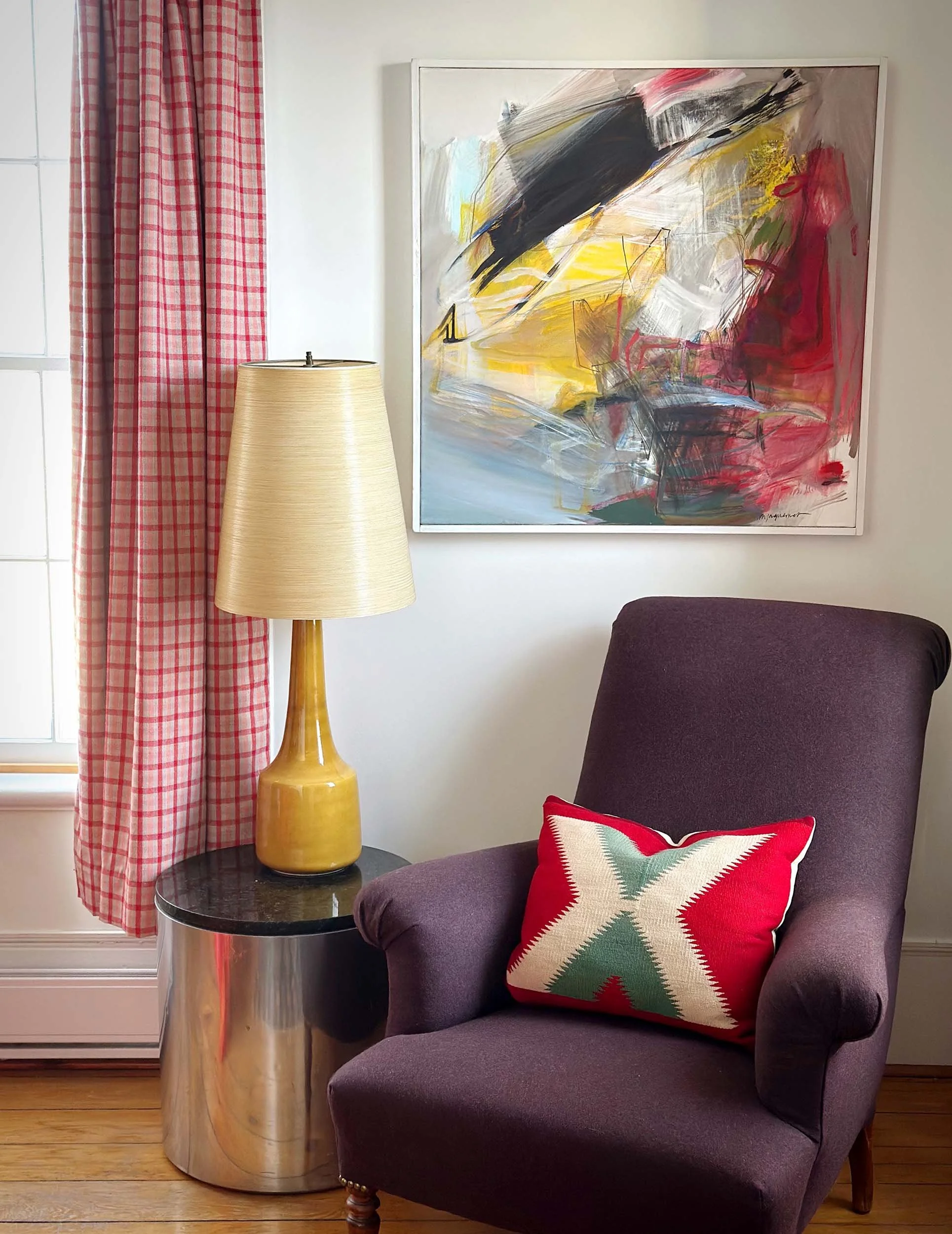 A purple armchair with a red, green, and white patterned pillow, a side table with a yellow vase lamp, a large abstract painting with black, yellow, red, and white brushstrokes on the wall, and red checkered curtains by a window.