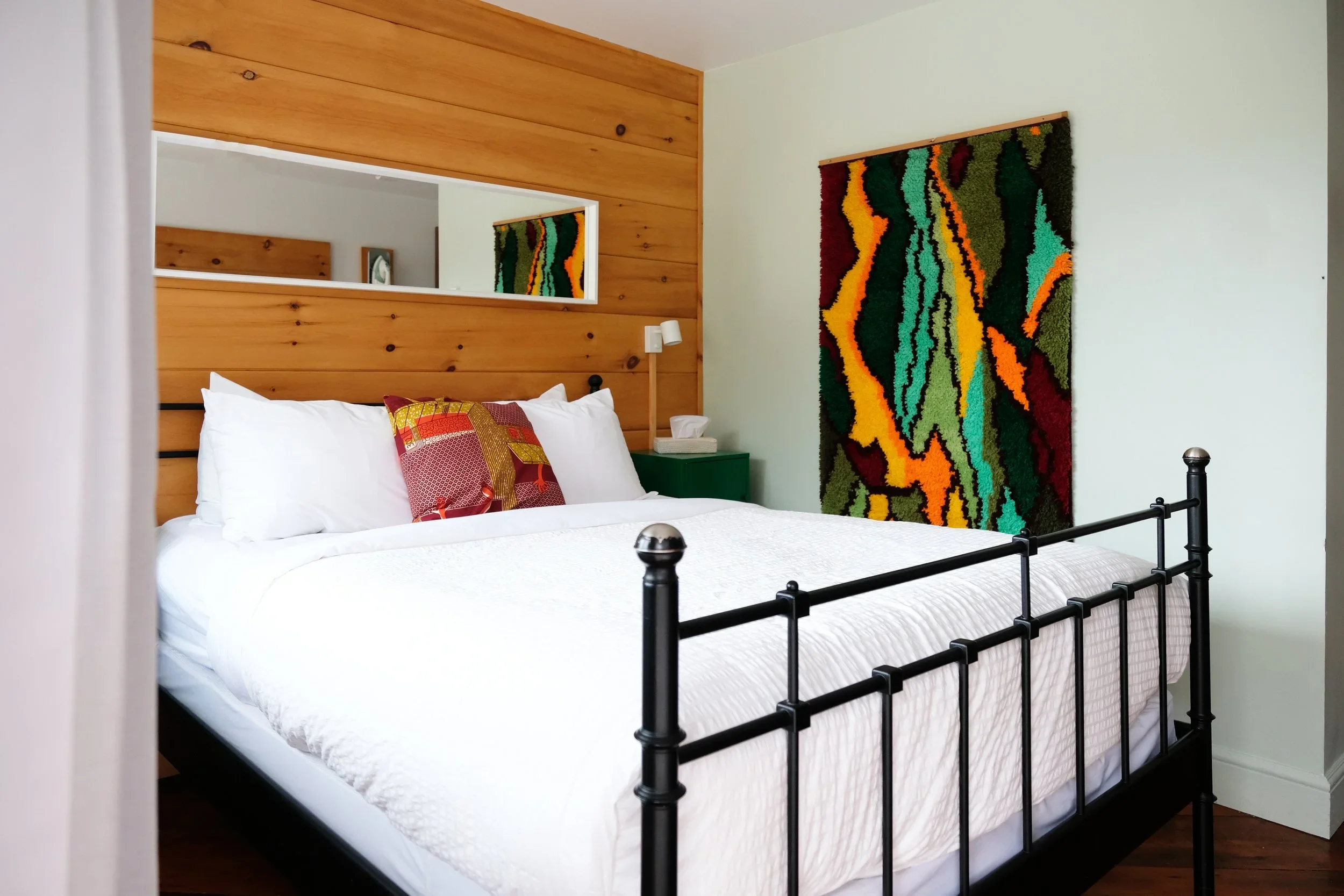 A hotel room with a black metal bed frame, white bedding, and a cushion with a red and gold patterned cover. The wall behind the bed features a wooden panel and a colorful abstract textile art piece. A white mirror is mounted on the wall, reflecting a partially visible clock and the room's ceiling. A small green nightstand with tissue box and a white wall-mounted reading light are beside the bed.