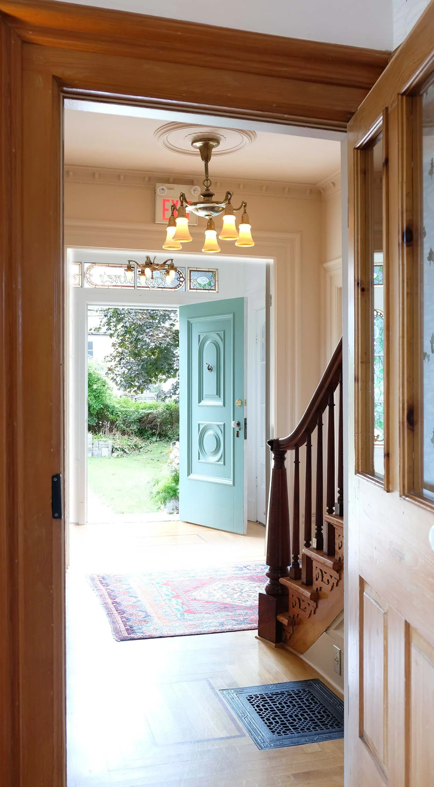 View of an entryway with open front door, leading outside to a garden, with a staircase on the right, a chandelier overhead, and a patterned rug on the wooden floor.
