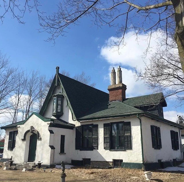 A large white house with black shutters and a steep dark roof, surrounded by leafless trees under a partly cloudy sky.