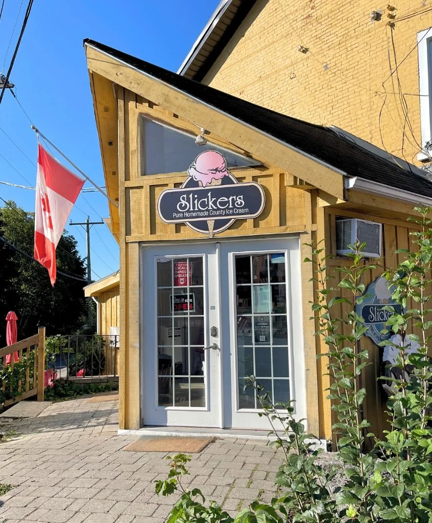 Front of Slickers ice cream shop with a sign featuring a pink ice cream cone mascot, a Canadian flag to the left, and a glass double door entrance.