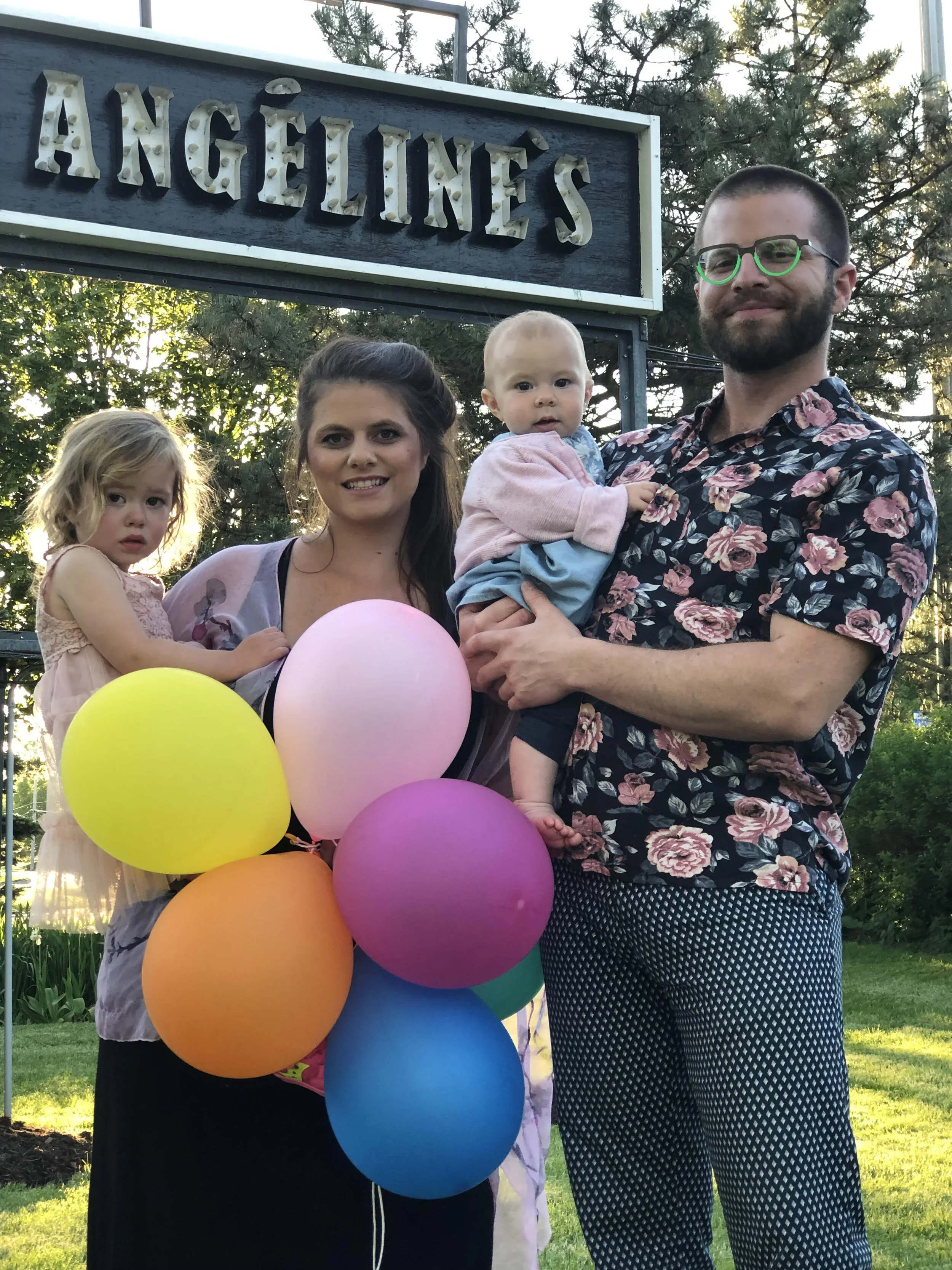 Group of four people, including two young children and two adults, standing outdoors in front of a sign that reads "Angeline's" with lightbulb adornments, holding colourful balloons, with trees and sunlight in the background.