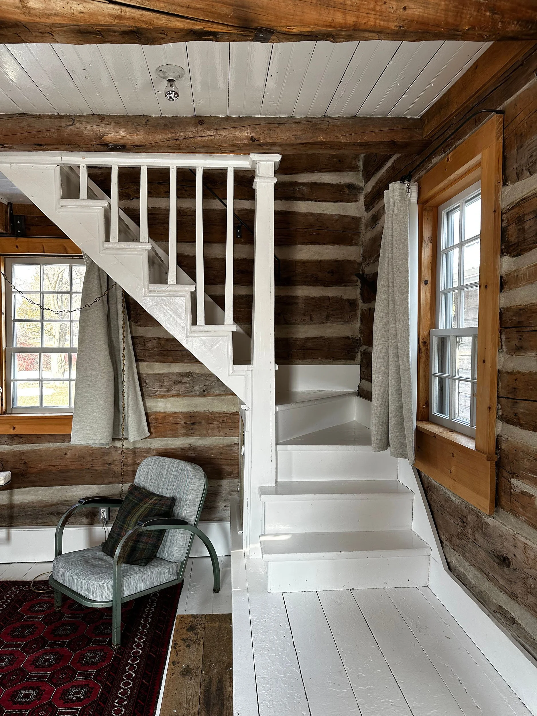 Interior of a rustic wooden house with exposed logs, a white staircase, a window with curtains, a chair with a pillow, and a red patterned rug.