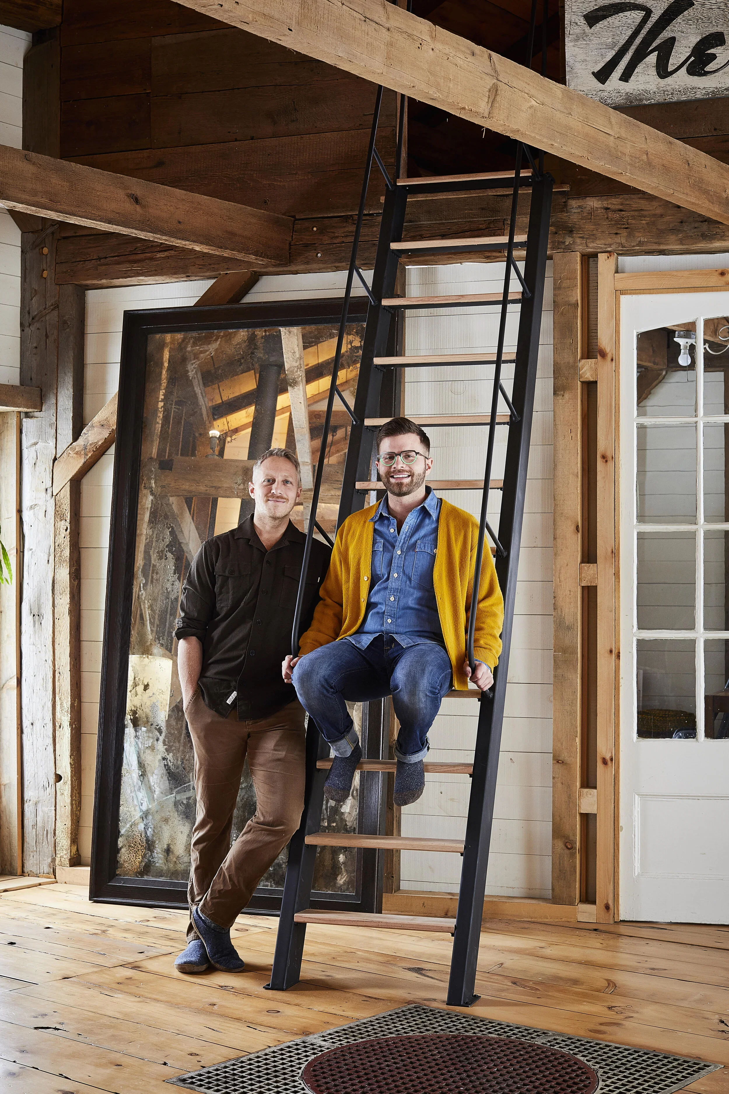 Two men inside a rustic wooden room with a ladder. One man is sitting on the ladder, smiling, wearing glasses and a yellow cardigan. The other man is standing beside him, smiling, with a black shirt and light brown pants.