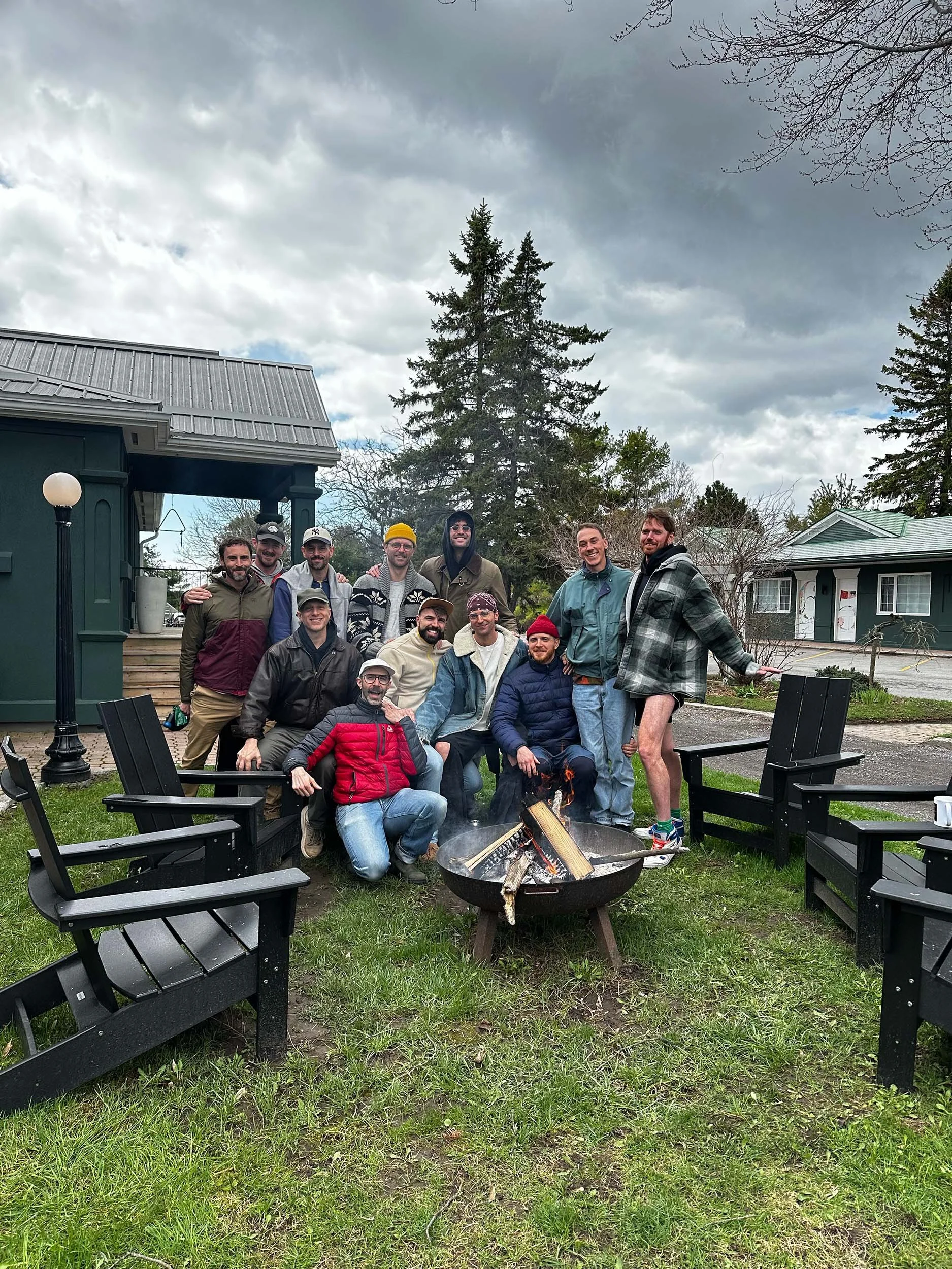 A group of people posing outdoors near a fire pit with wood burning, surrounded by four black patio chairs, with buildings and large trees in the background on an overcast day.
