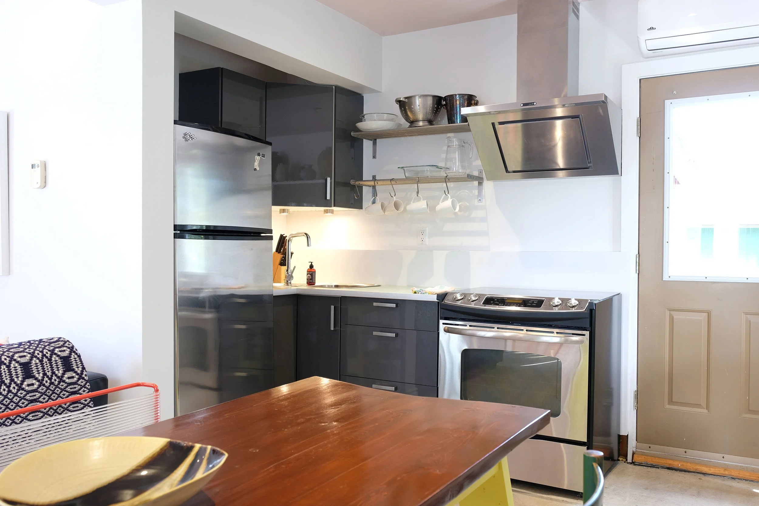 A modern kitchen with stainless steel appliances, dark cabinets, open shelves with bowls and cups, and a dining table with a decorative bowl in the foreground.