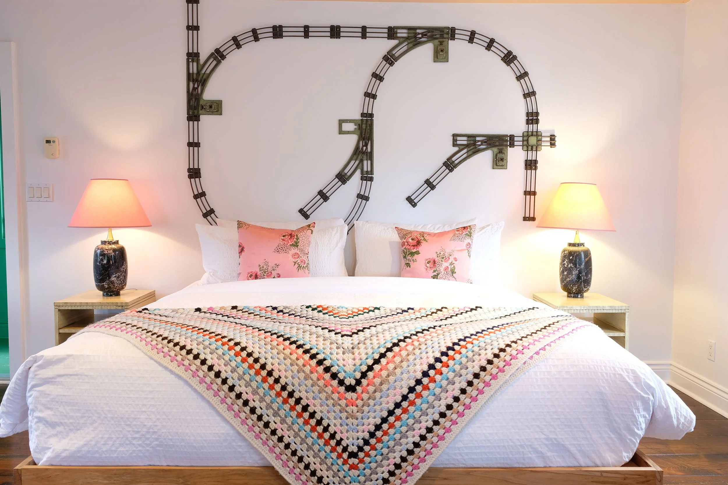 A bedroom with a decorated bed featuring white bedding, floral pillows, and a multicolored crocheted blanket. There are two matching black marble nightstands with pink lamps. The wall has a decorative metal clock in a vintage train design.