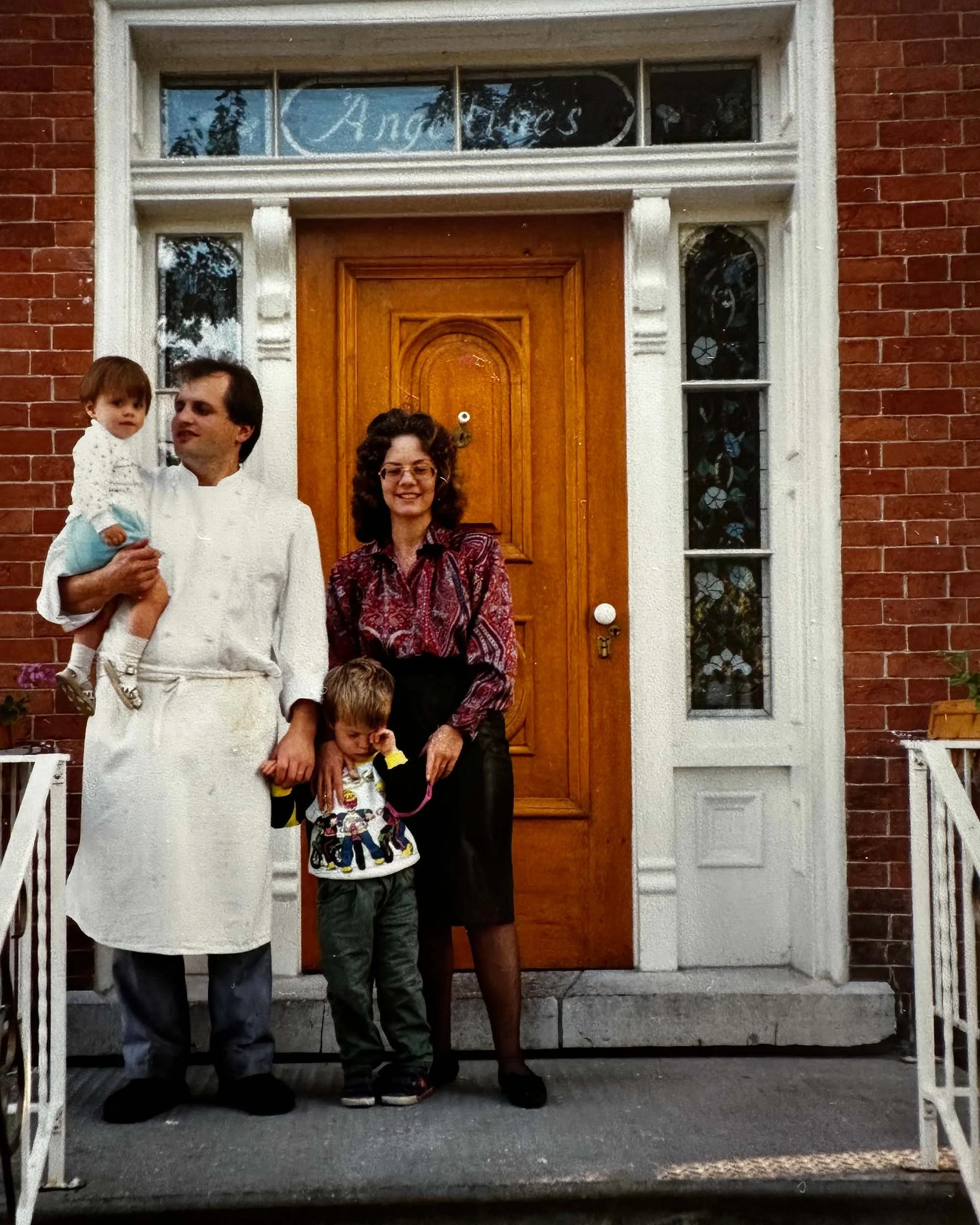 A man holding a young girl, and a woman standing in front of a brick house with a wooden door and decorative stained glass windows. The man and girl are on the left, the woman on the right, and an additional child's head is visible in the lower part of the image.