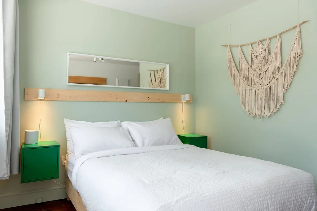 A cozy bedroom with a light green wall, a large mirror above the bed, and a macrame wall hanging decoration. The bed has white bedding and two green side tables with lamps.