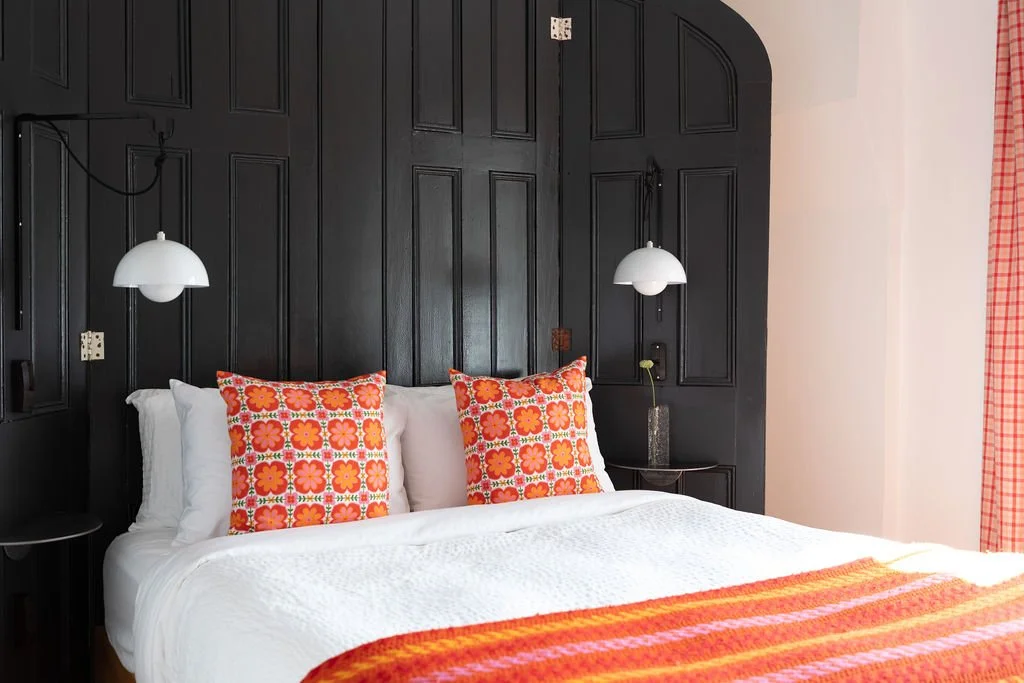 A neatly made bed with white bedding and two decorative pillows with orange and red floral patterns. The bed is against a black wooden headboard and has two white hanging pendant lights, one on each side. There is a small nightstand with a glass vase and single flower on the right side, and patterned curtains on the window to the right.