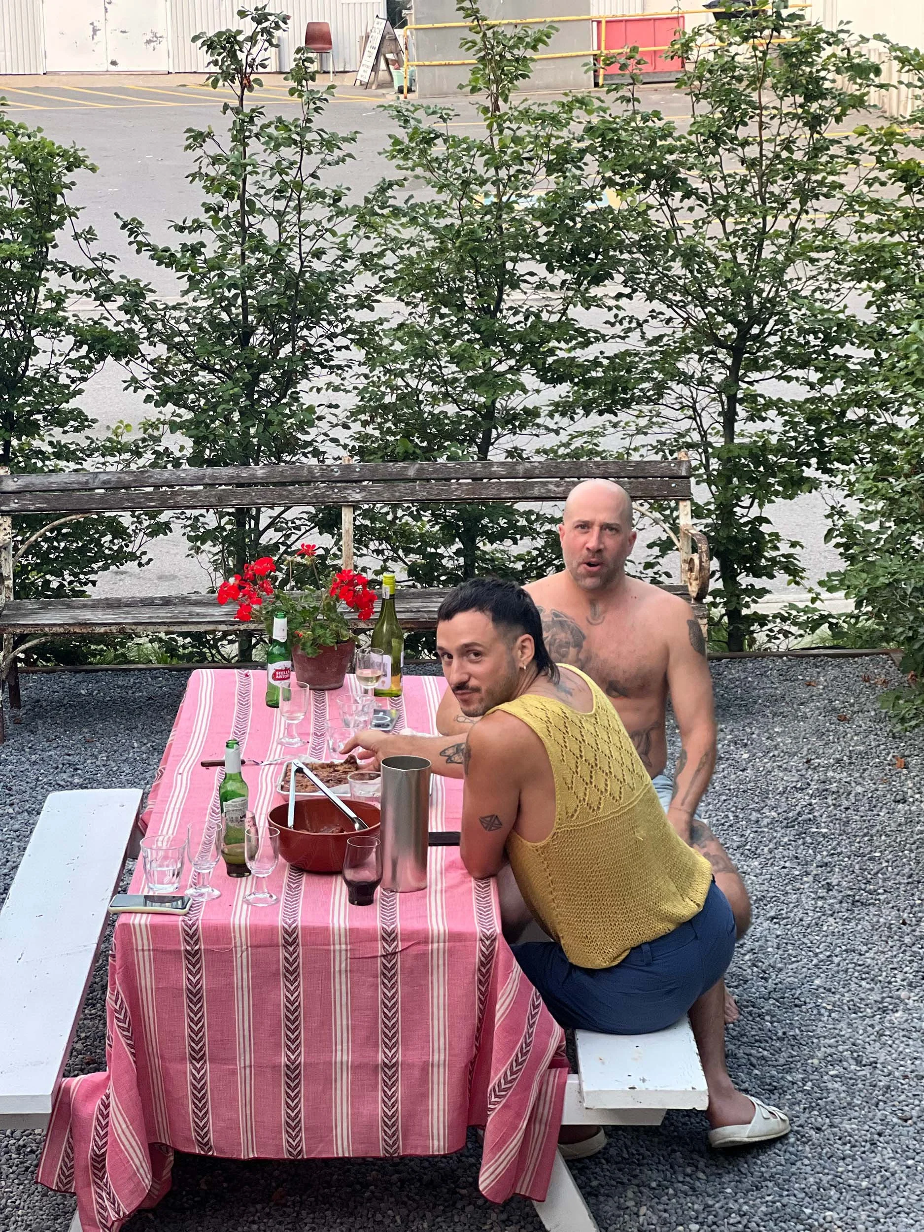 Two shirtless men at an outdoor gathering. One is sitting at a table with a pink striped tablecloth, and the other is kneeling beside him. The table has bottles, glasses, and a potted plant with red flowers. The background features a wooden bench, some greenery, and a gravel ground.