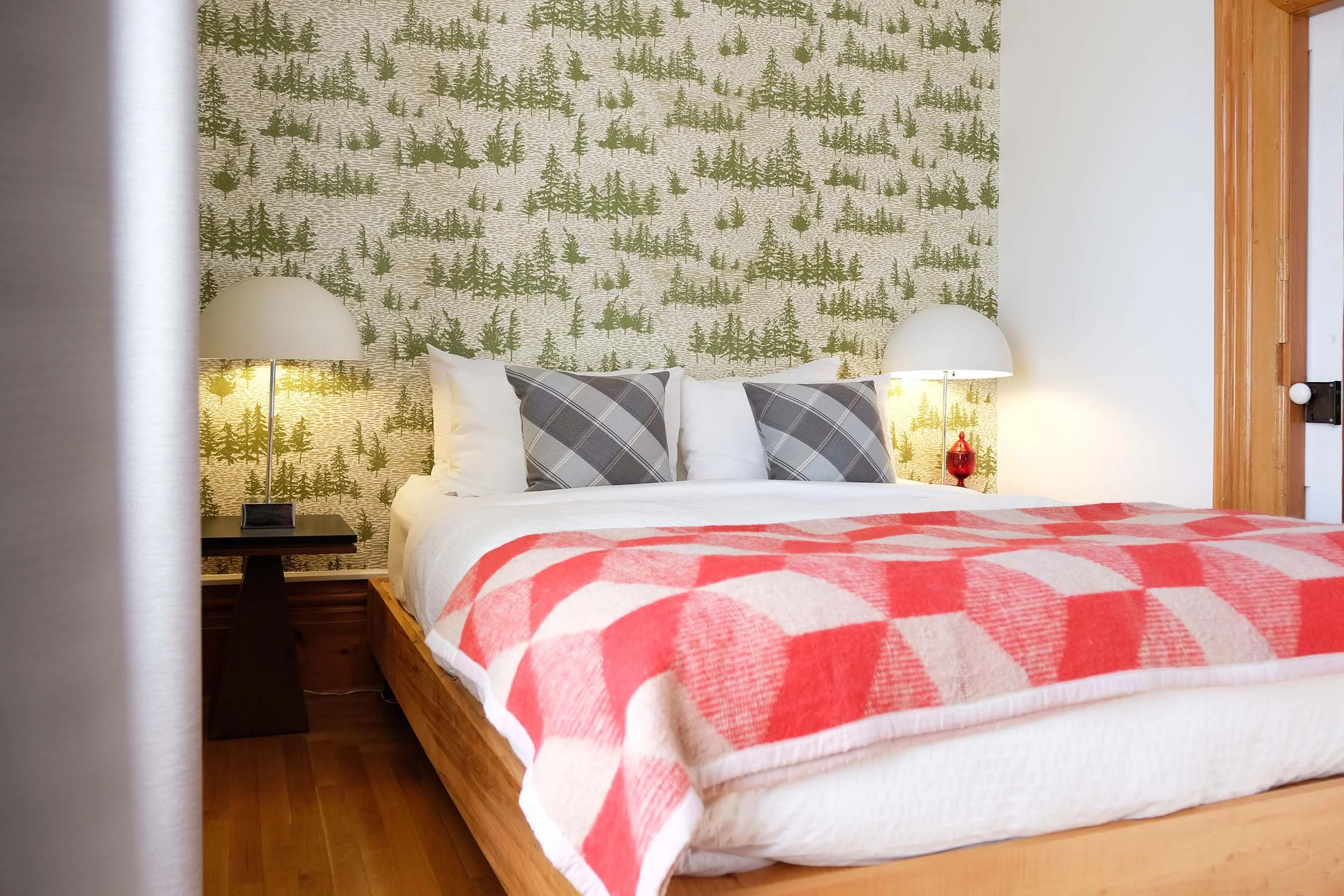 A cozy bedroom with a wooden bed frame, white bedding, and a red and white checkered blanket. There are two gray and white plaid pillows and two white pillows against a wall with forest-themed wallpaper. Two white bedside lamps with domed shades sit on black bedside tables. Part of a wooden doorframe is visible on the right.