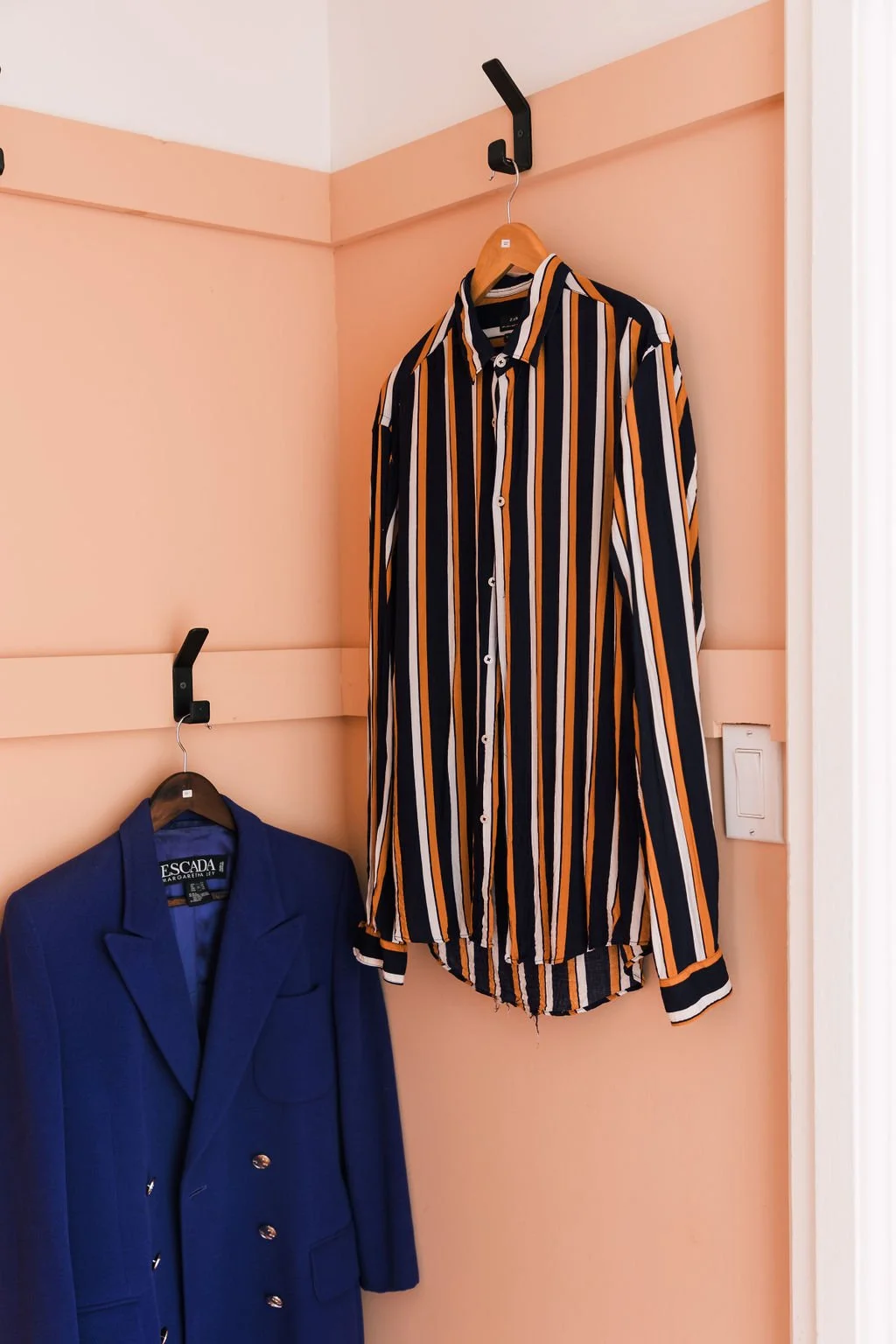 A peach-colored wall with two black hooks holding clothing: a striped dress shirt hanging on a wooden hanger and a blue blazer on a black hanger.