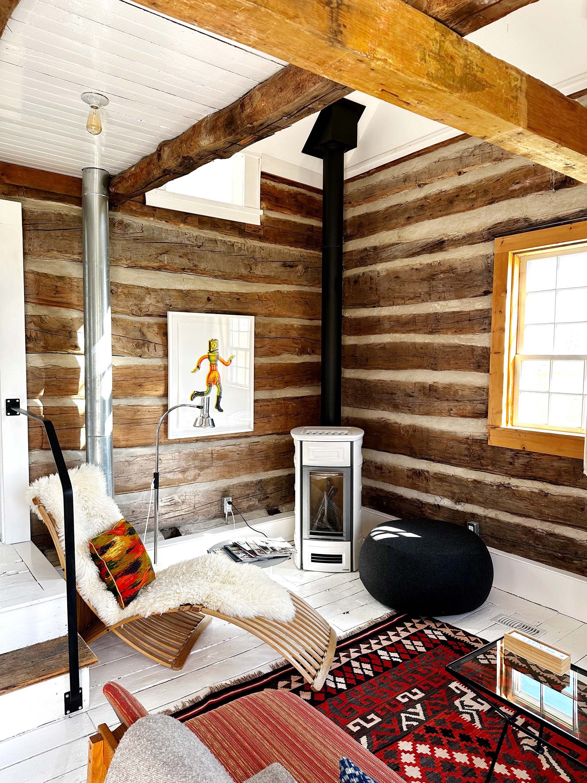 Cozy living room with wooden beams, white-painted floor and walls, a window with wooden trim, a black round pouf, a small white wood stove, a colorful floor rug, a cane lounge chair with a plush cushion and colorful pillow, a framed art piece on the wall, and a glass-top side table.