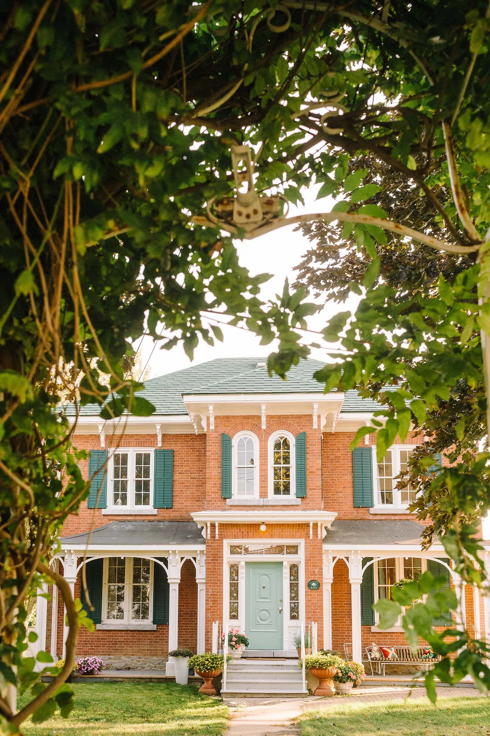 Front view of a three-story brick house framed by green leafy trees with a porch, flower pots, and a mint green front door.
