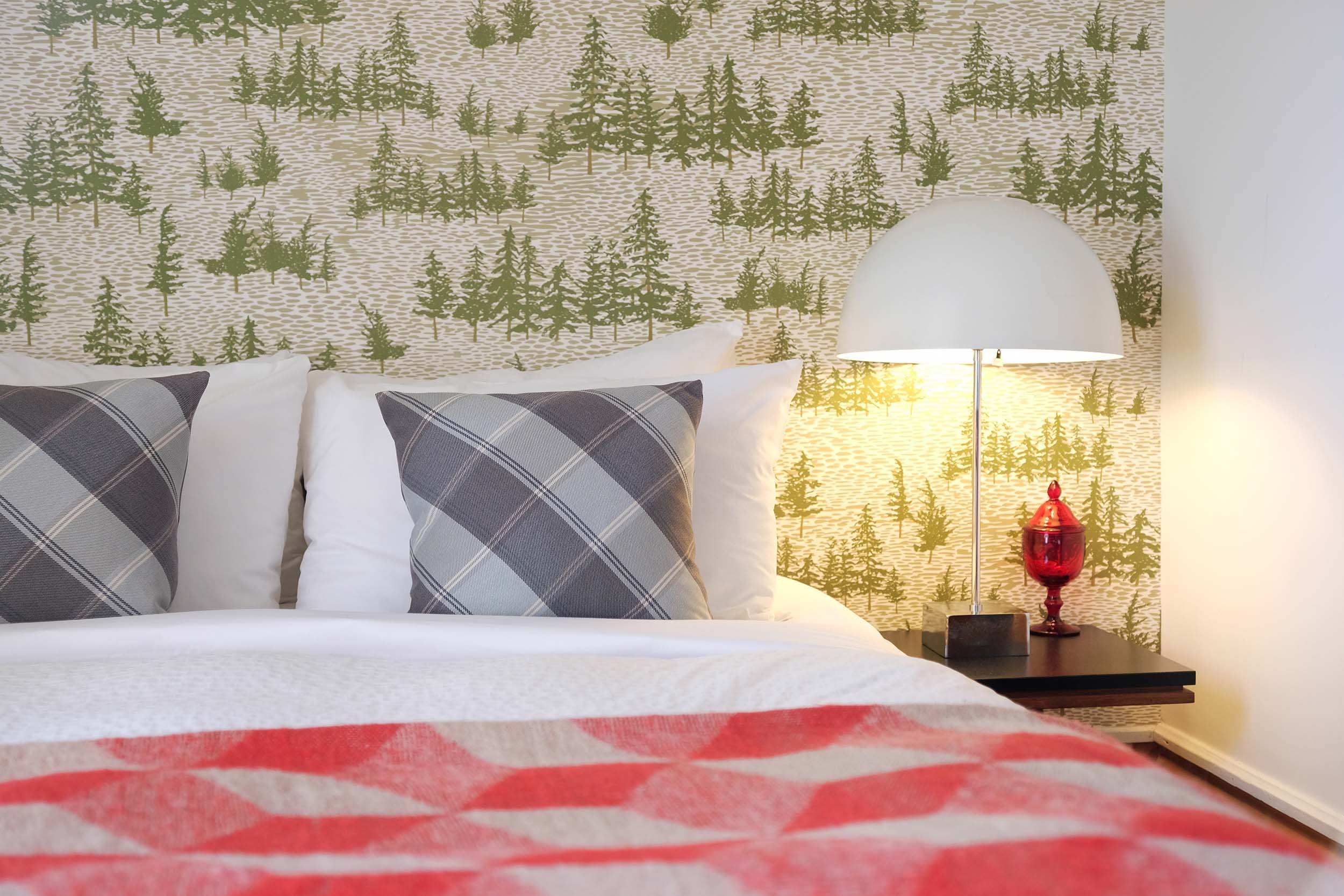 A bed with plaid pillows, a bedside lamp, and a red decorative vase in front of a wallpaper with a forest pattern.