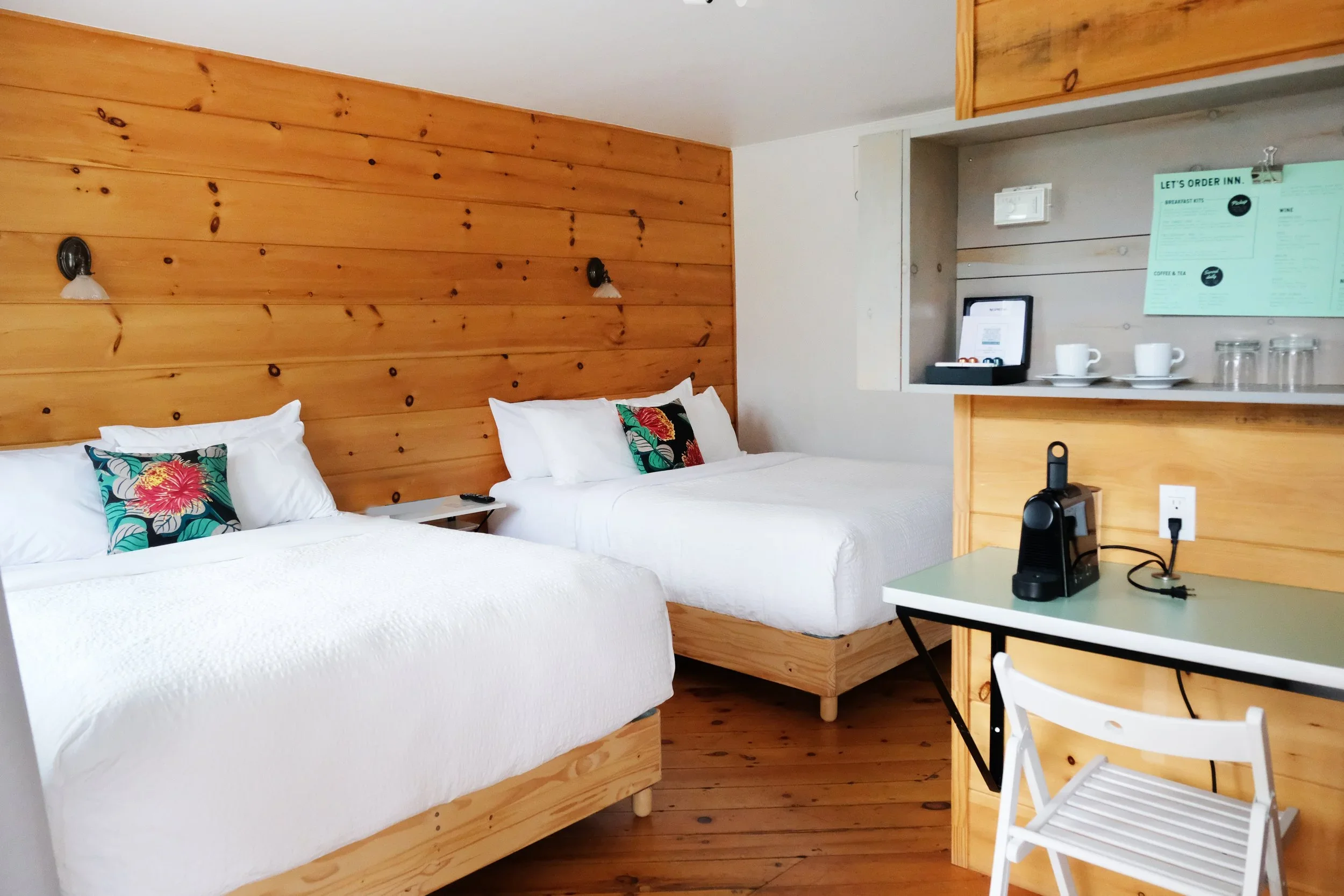 Hotel room with two queen beds with white bedding and colorful floral pillows, wooden wall paneling, wall-mounted lamps, small bedside tables, a corner with a framed tablet, on the right a kitchenette with cups, glasses, and a menu, and a small white table with a chair and electronic devices.