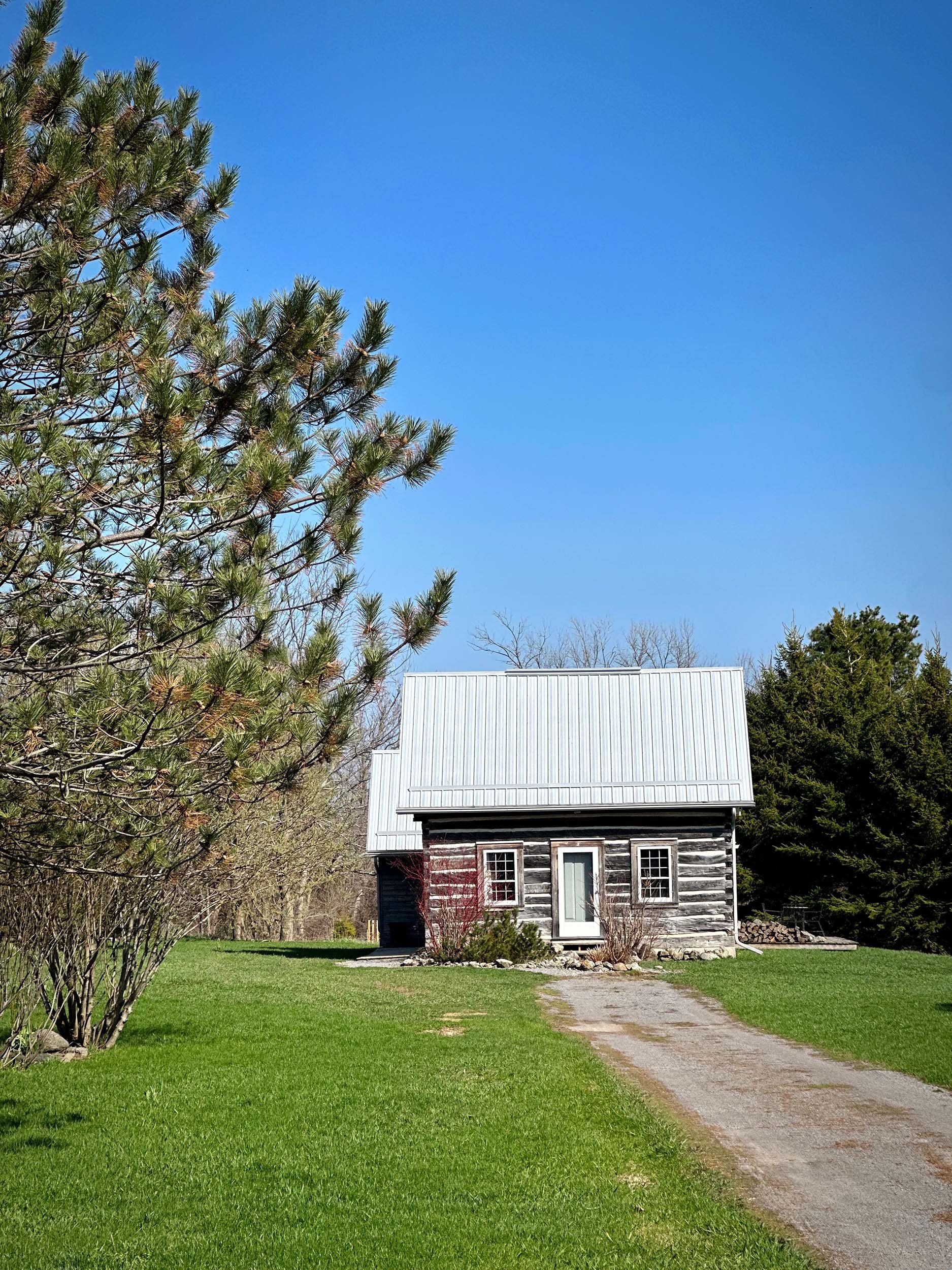 A small, old wooden house with a metal roof, located on a grassy area under a clear blue sky. There are trees around the house, and a gravel pathway leads to its door.