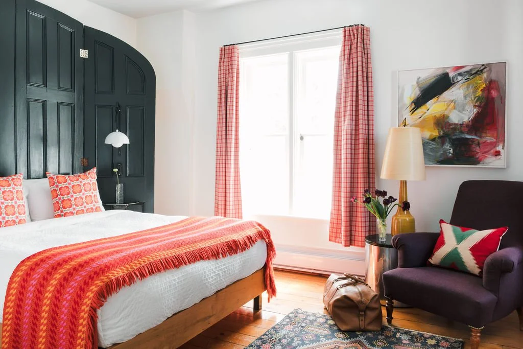 A cozy bedroom with a black headboard, white bedding, and red-orange patterned pillows and throw blanket. There are pink checkered curtains, abstract art on the wall, a purple armchair with a star-patterned pillow, a yellow lamp, a small side table with flowers, a leather bag on the floor, and a window letting in natural light.