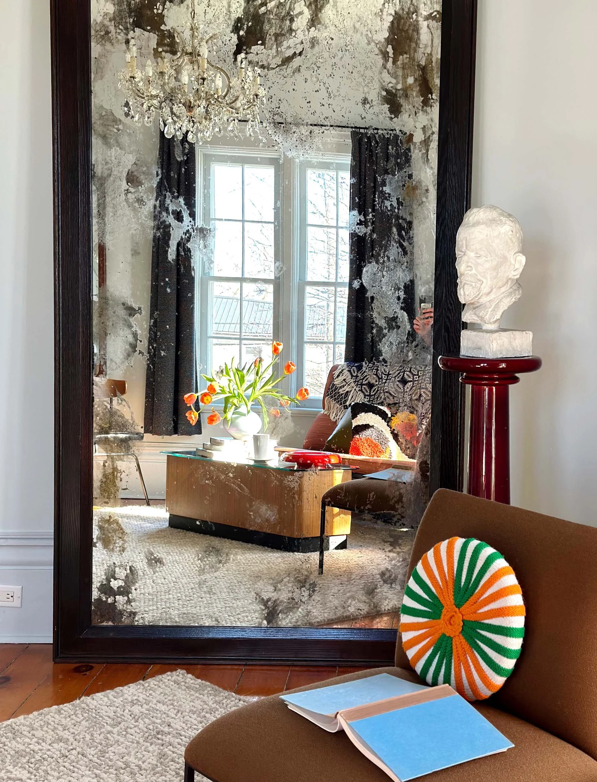 A large mirror reflecting a room with a crystal chandelier, window with black and white curtains, and a white bust sculpture on a red pedestal. The room has a beige carpet, a round wooden table with a vase of orange tulips, and a brown chair with an open notebook and a blue book on it. A person is taking the photo, visible in the mirror's reflection.