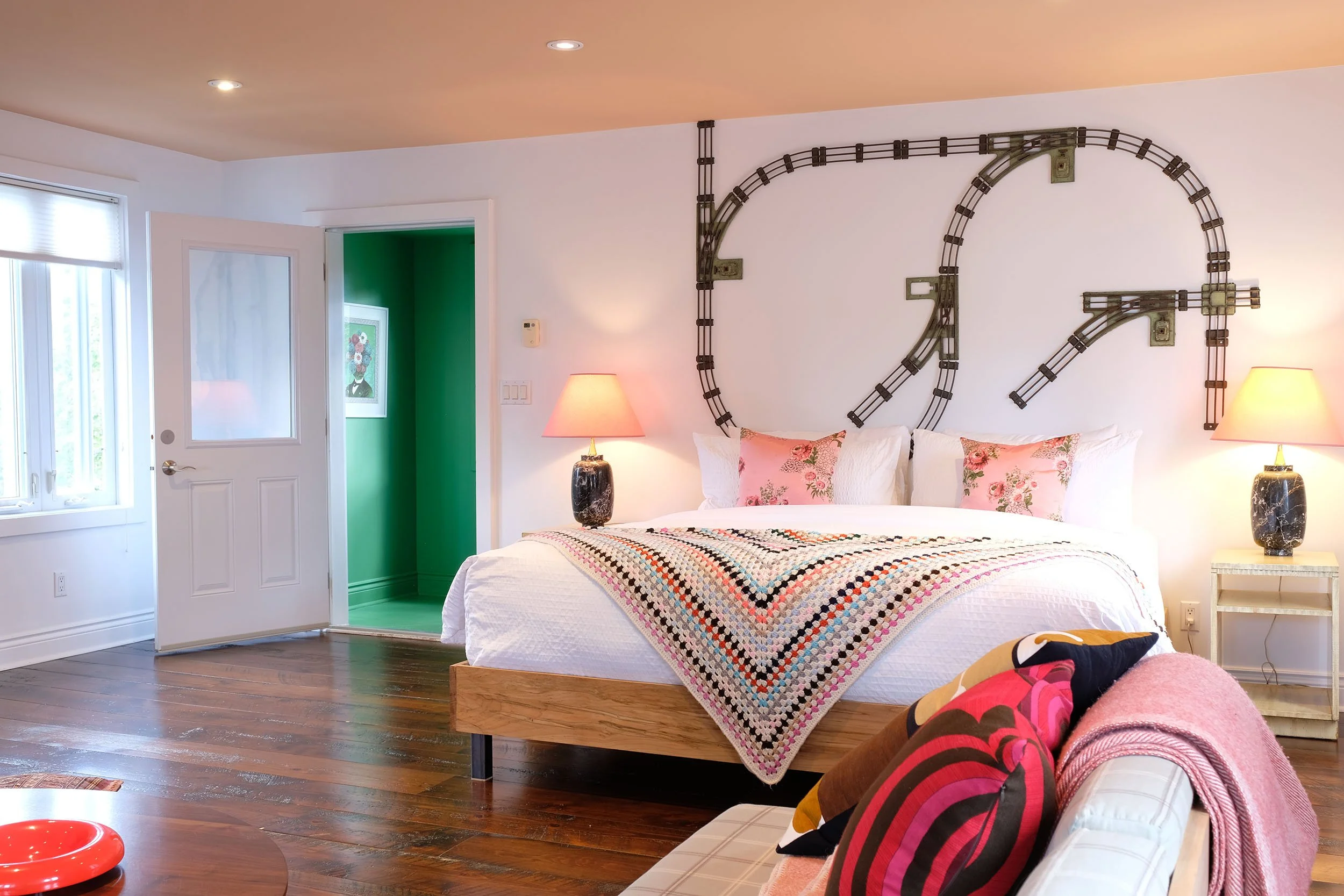 A bedroom with a bed, nightstands with lamps, a crochet blanket, and a colorful blanket on a nearby sofa. The wall features large metal artwork resembling a clock without hands. There's a partially open door showing a green room with framed art. The room has hardwood floors, white walls, and natural and artificial lighting.