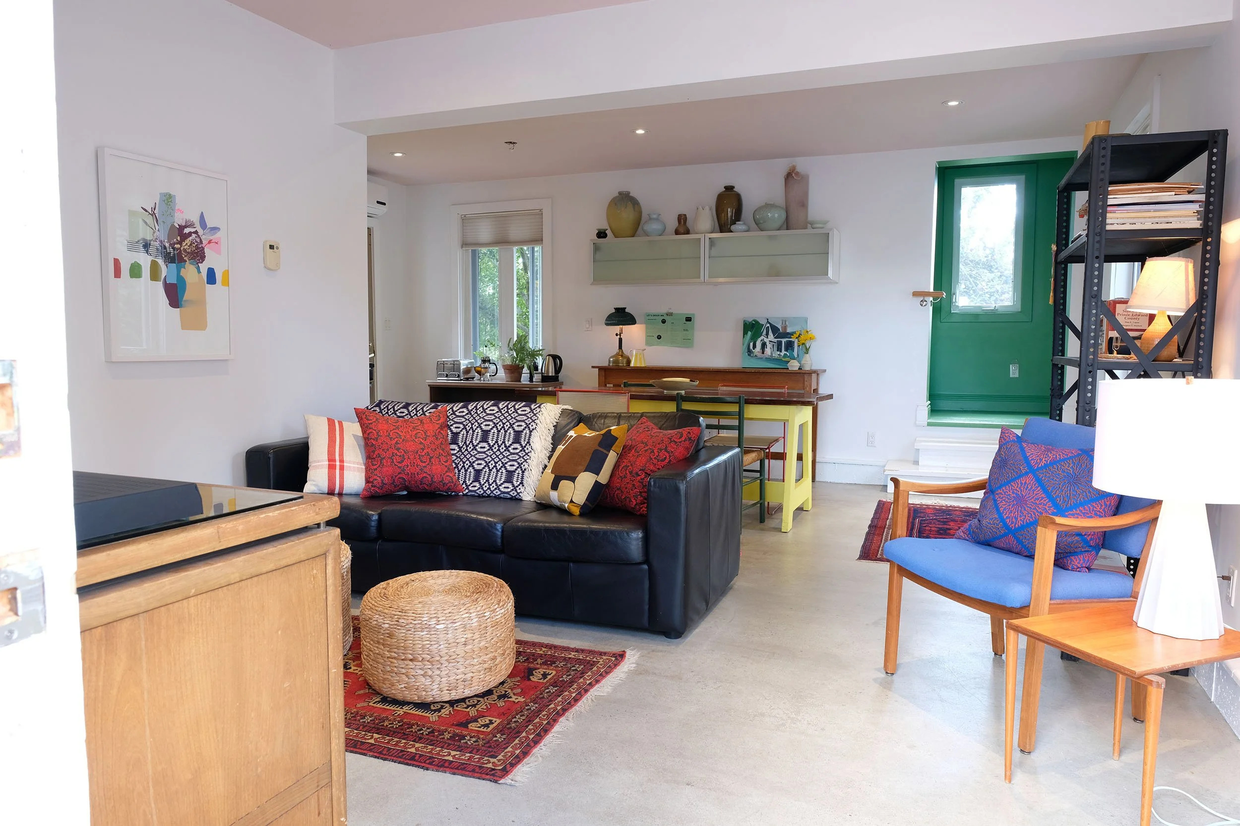 A cozy living room with a black sofa adorned with colorful pillows, a wooden coffee table, and a red patterned rug. The room features a variety of decorative items, including vases on a white shelf, a painting on the wall, and a bookshelf with books. There are two chairs, one of which is blue with a patterned pillow, and the room has a bright atmosphere with natural light coming through a window with a green frame.