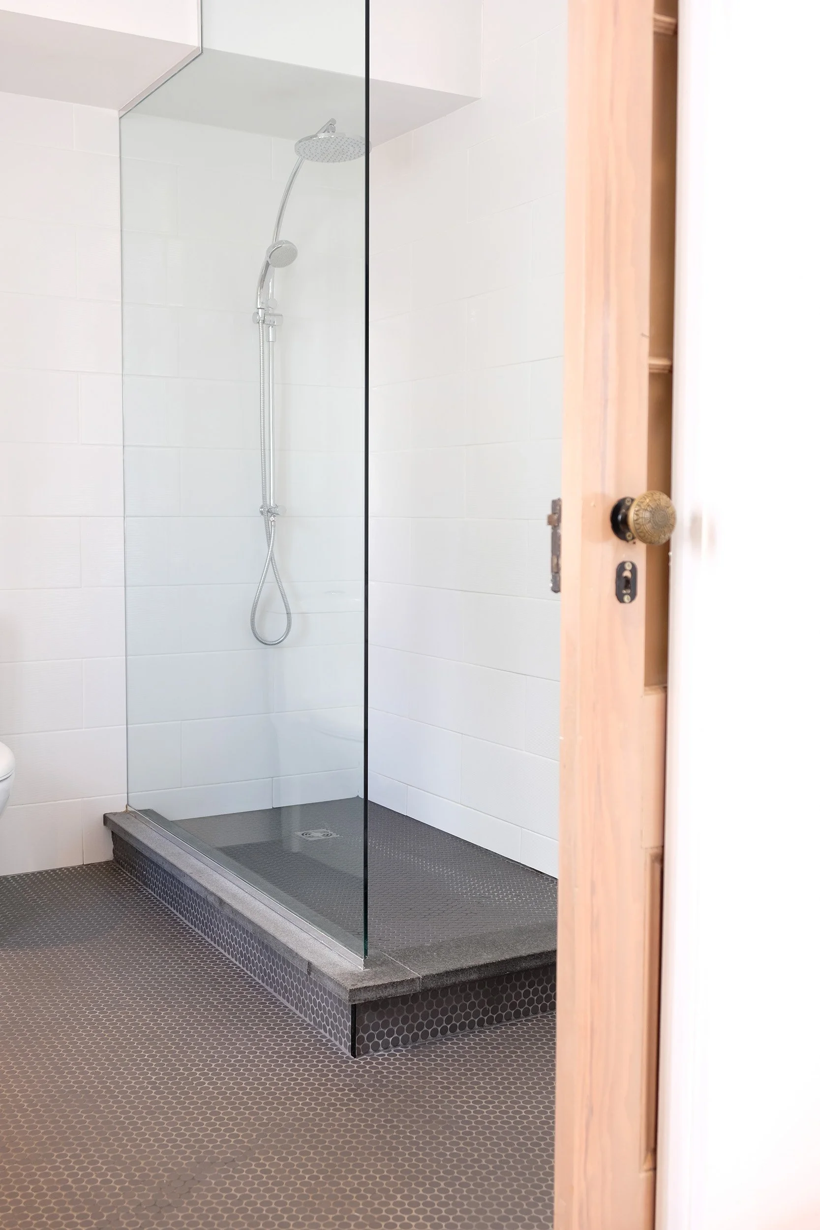 A modern, minimalist shower with white tiled walls, a silver adjustable showerhead, and a glass door. The shower floor is dark gray with small hexagonal tiles, and it sits on a slightly elevated platform. Part of a light wooden door with a vintage-style round brass doorknob is visible on the right side of the image.