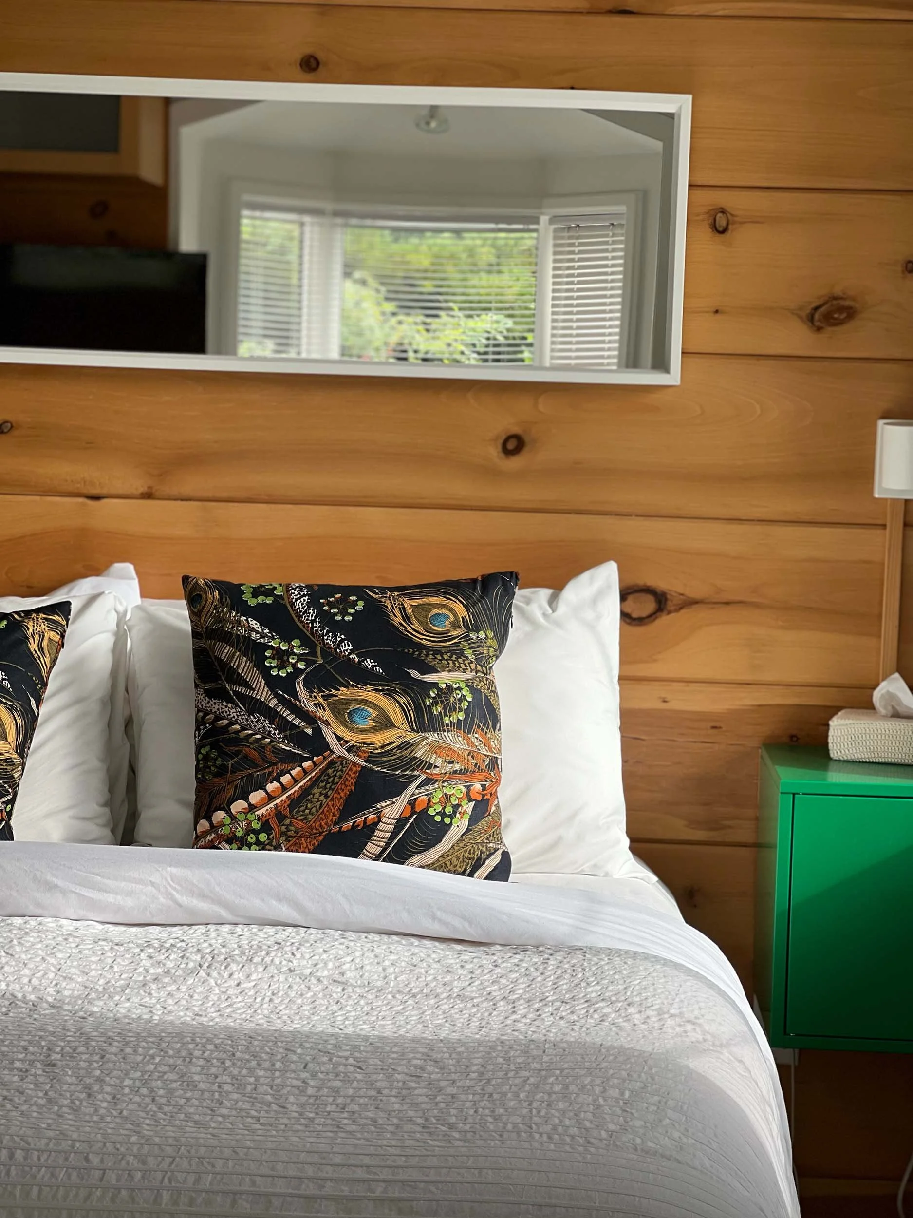 A mirror on a wood-paneled wall reflecting a window with blinds and greenery outside. Part of a white bed with floral pillows and a green nightstand with a white lamp are visible.