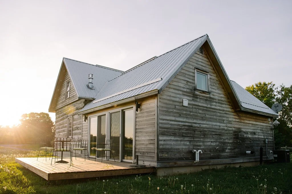 A rustic wooden house with a metal roof, a small porch with chairs, and a grassy yard at sunset.