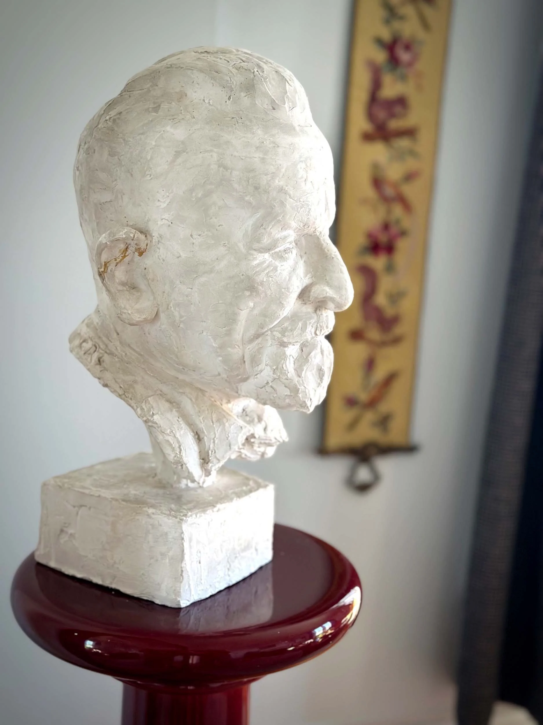 White bust sculpture of a man with detailed facial features, mounted on a white rectangular base, displayed on a red pedestal.