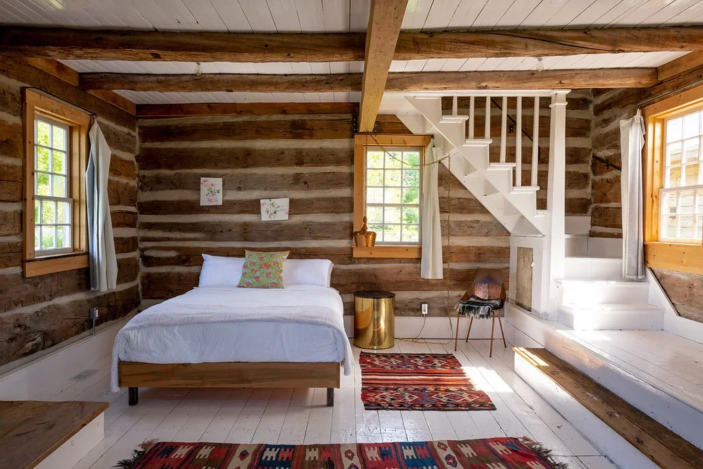A rustic bedroom with wooden log walls, white painted wood floors, and three windows with white curtains. There is a bed with white bedding and a colorful pillow, a small round side table with a gold finish, a patterned rug, a chair, and a staircase leading to an upper level.