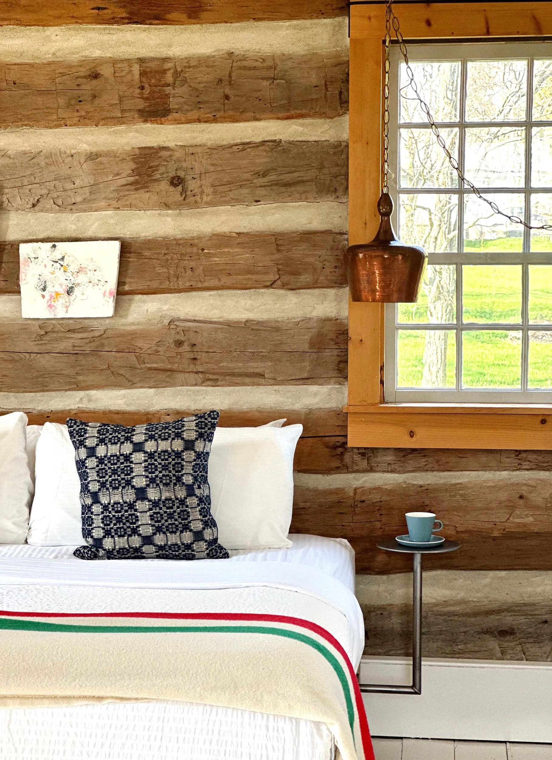 Interior of a rustic room with wood-paneled walls, a large window showing a view of trees and a grassy yard, a bed with white bedding and a navy patterned pillow, a small side table with a blue cup and saucer, a copper hanging lamp, and a small abstract painting on the wall.